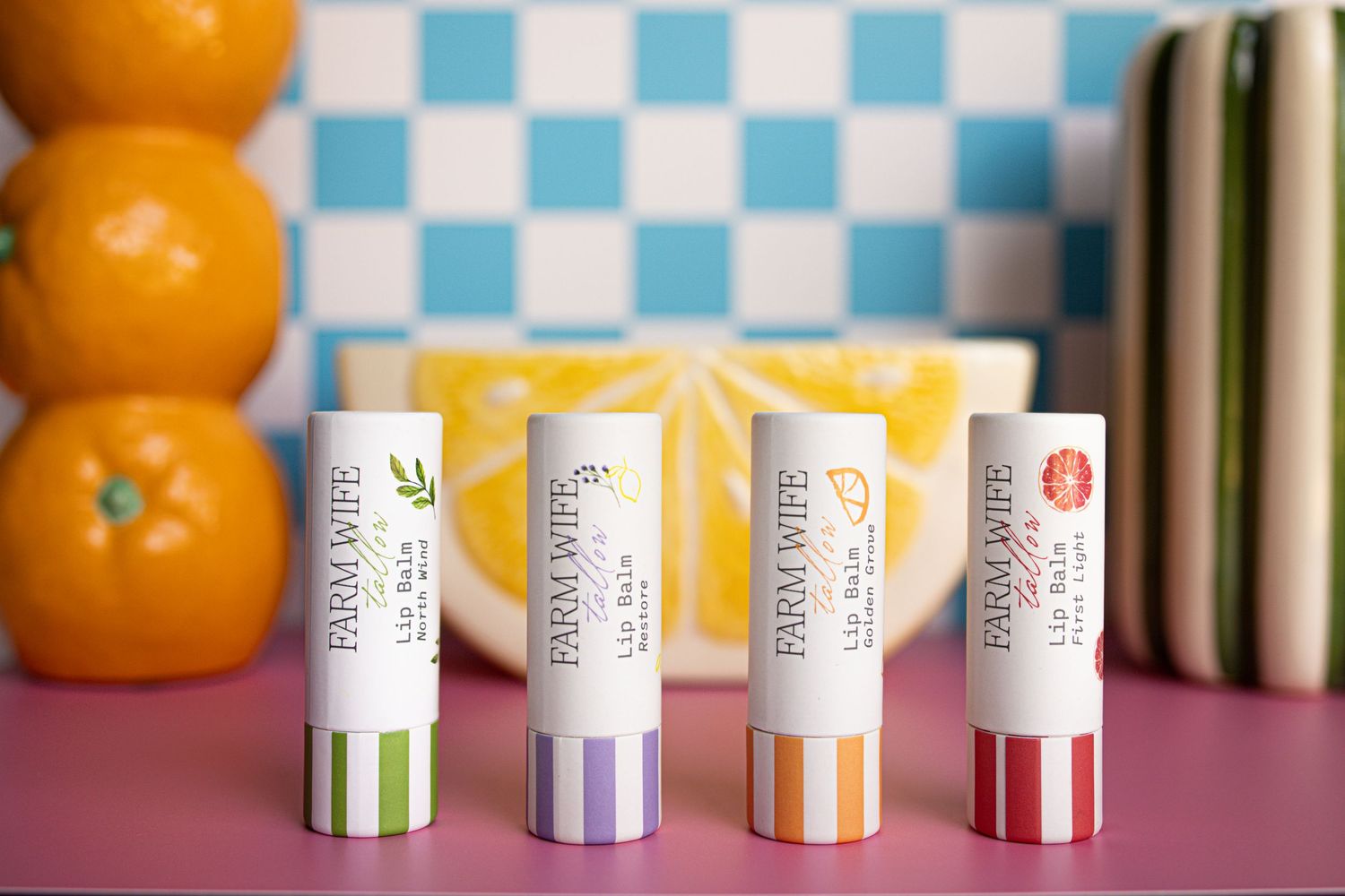 Four Farm Wife lip balm sticks in colorful striped packaging displayed on a pink surface with fruit and checkered backdrop.