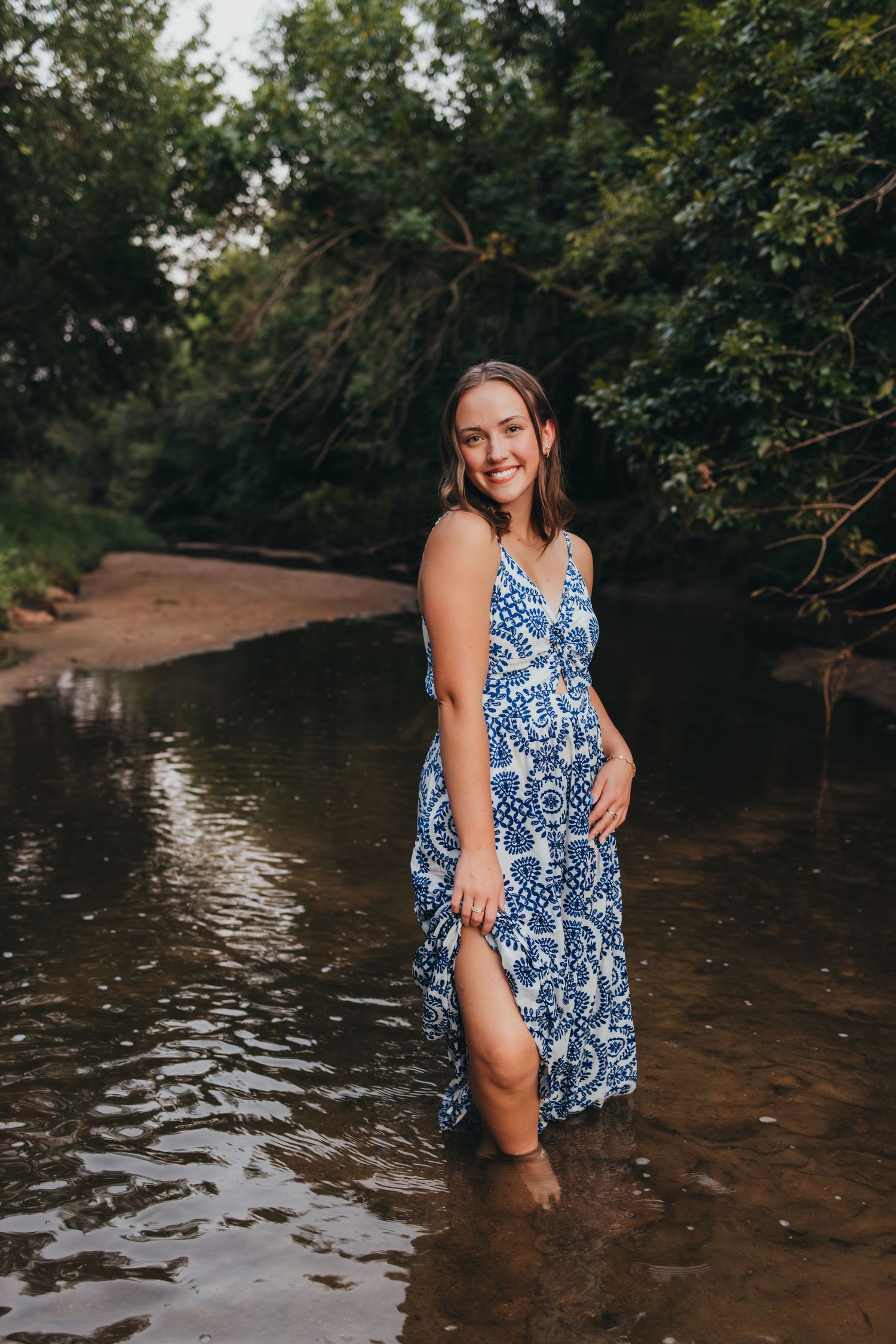 Someone wearing a blue and white patterned sundress poses playfully in shallow creek water on a summer afternoon.
