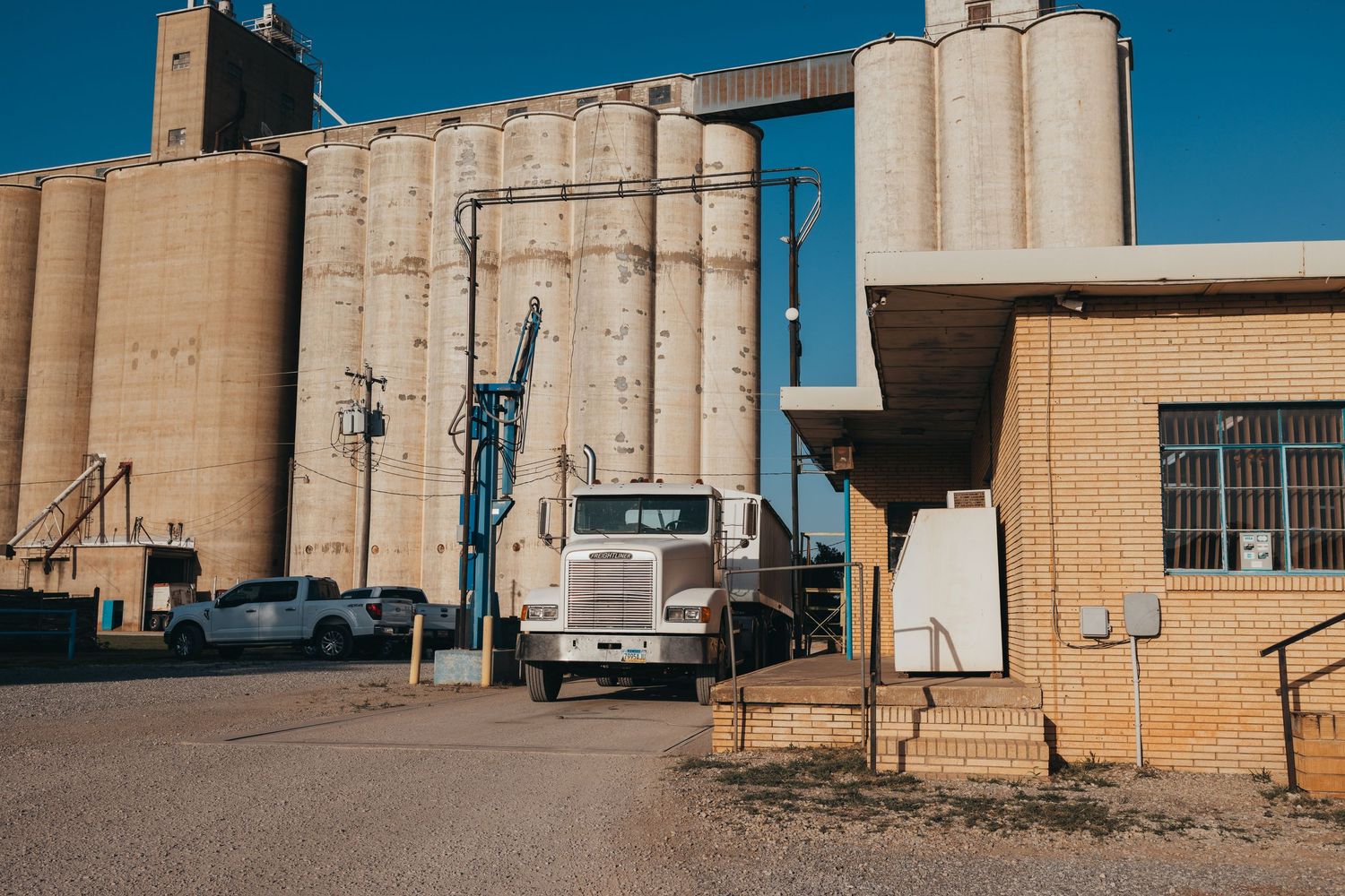 A white semi truck parked next to tall concrete grain silos under a bright blue sky.