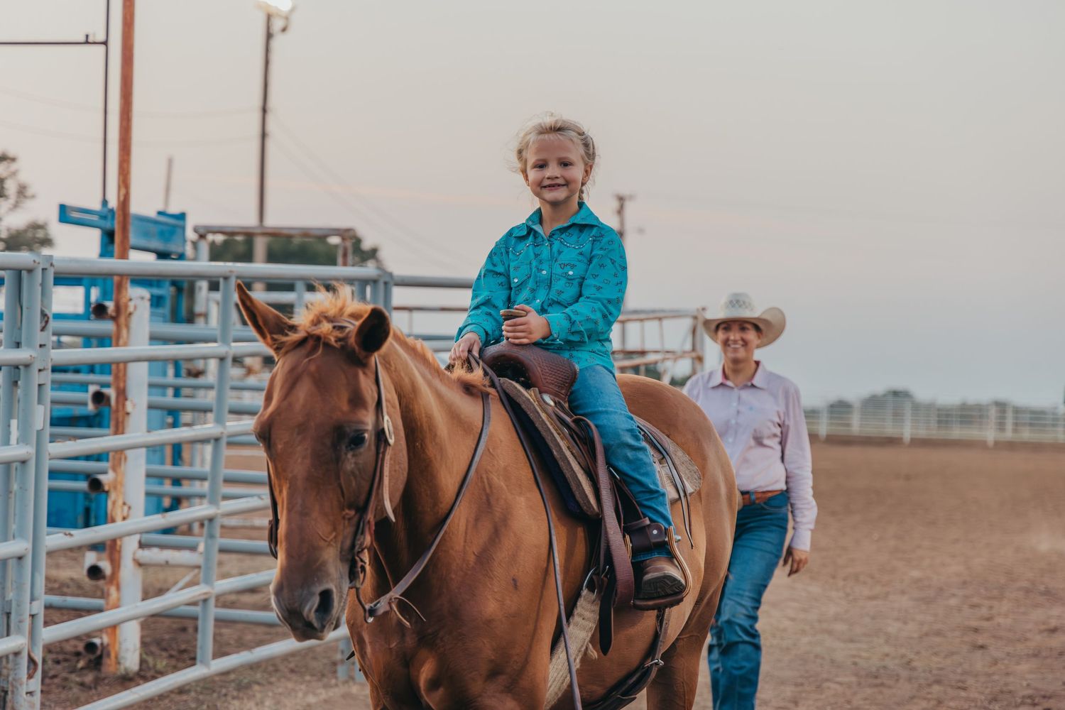 Young child in turquoise shirt riding a chestnut horse at a rodeo arena, guided by an adult in a white cowboy hat.