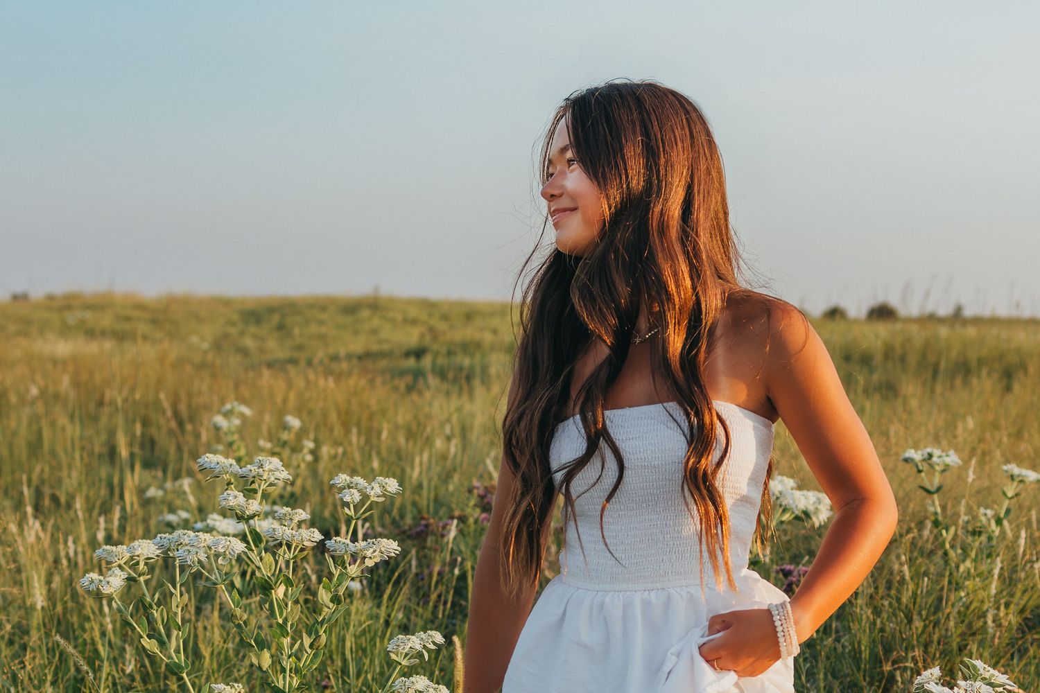 Woman in white dress walking through a sunny meadow with wildflowers and tall grass.