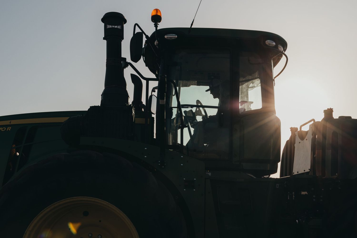 Silhouette of a John Deere tractor against a bright sunset sky on a farm field.