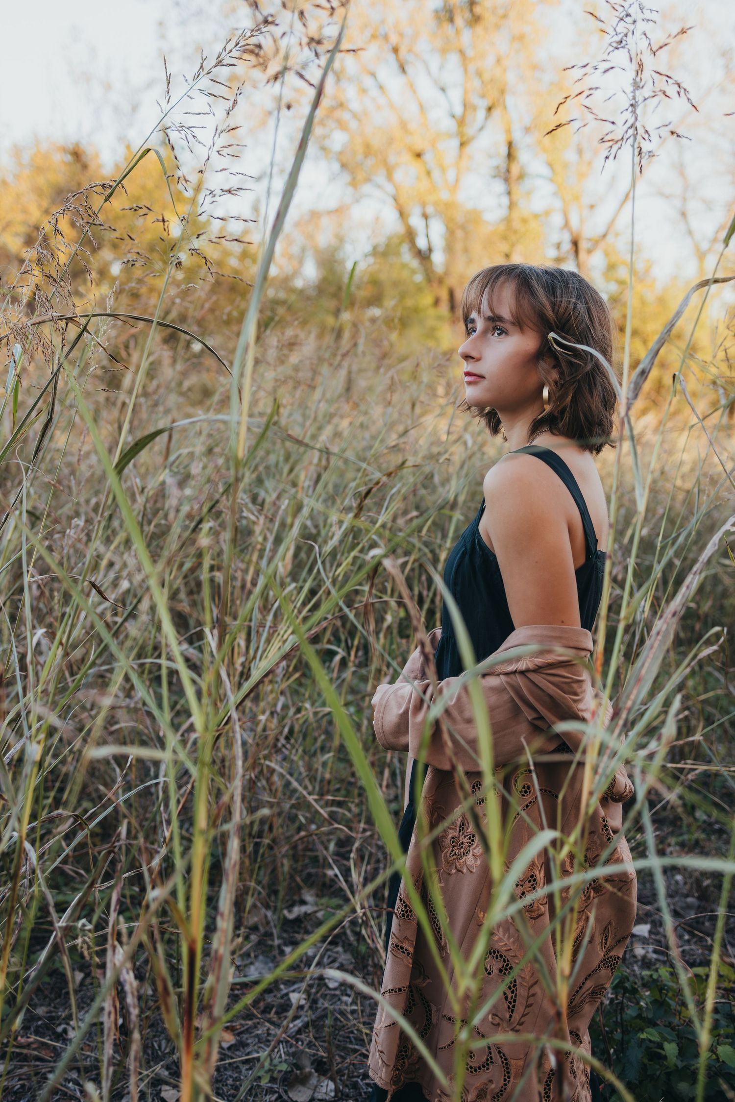 Person in dark top poses thoughtfully among tall golden grasses in autumn sunlight.