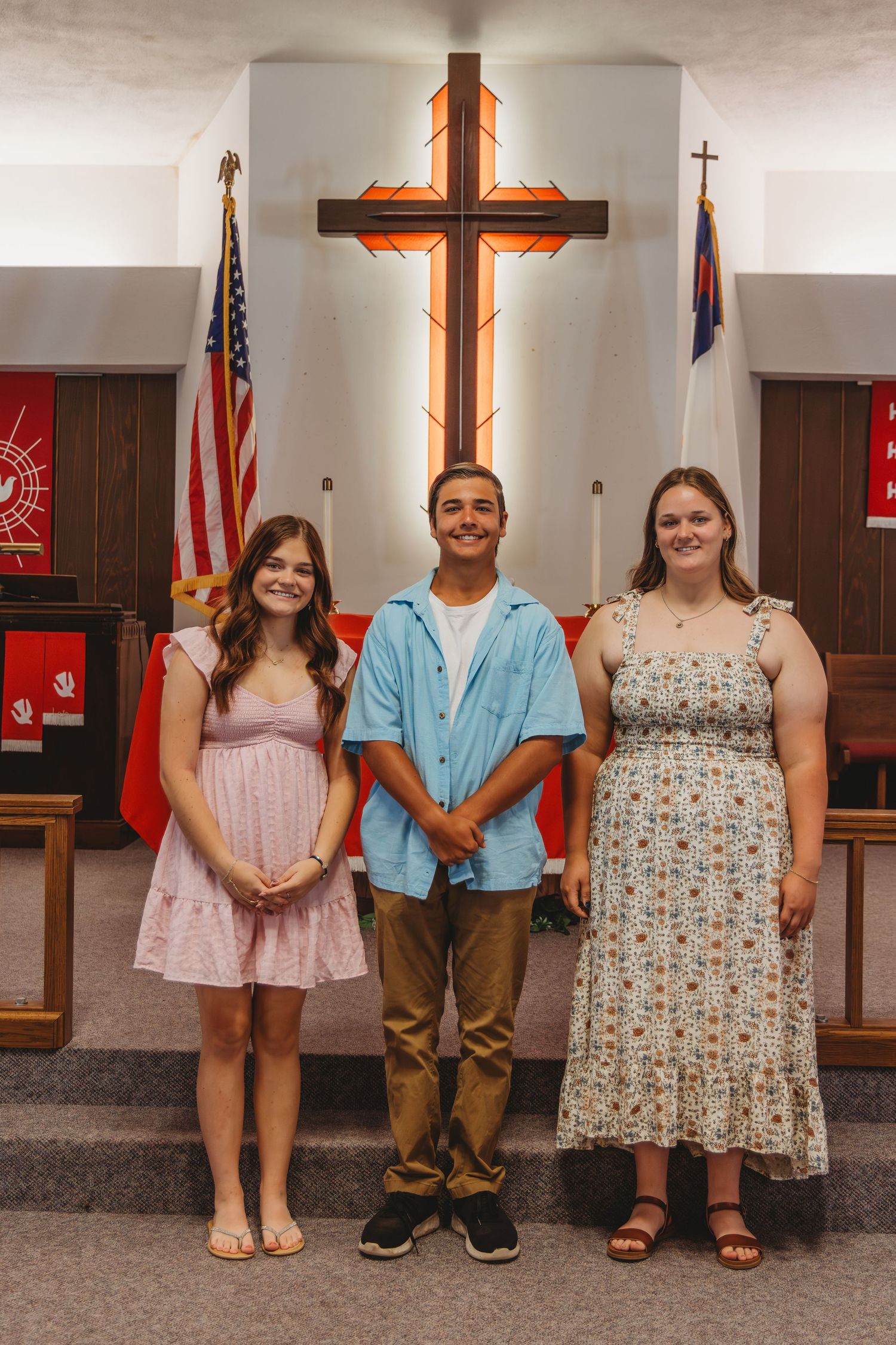 Three young people stand in front of a large illuminated wooden cross inside a church with American and Christian flags.