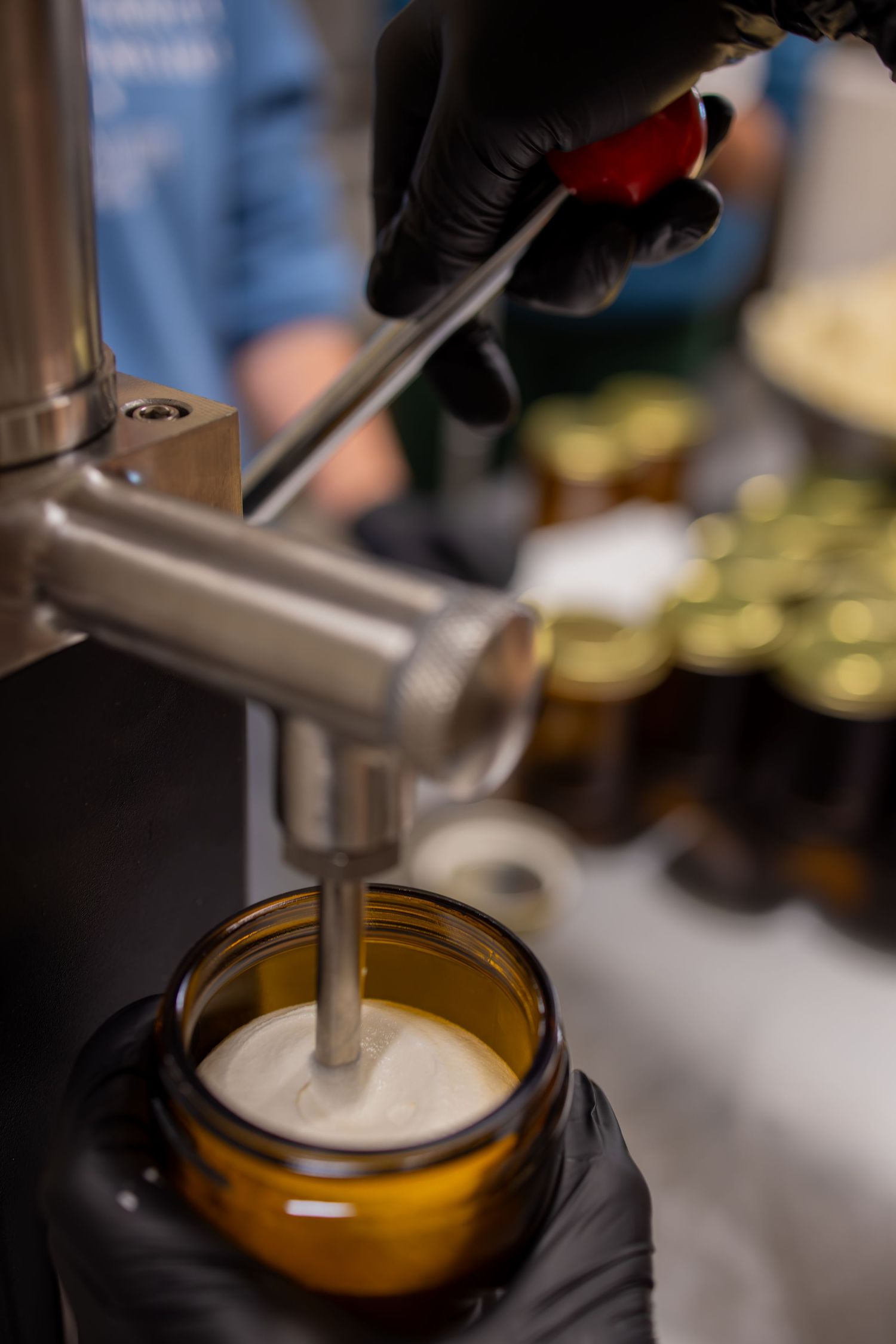 Close-up of a metal mixing equipment filling an amber jar with white cream product.