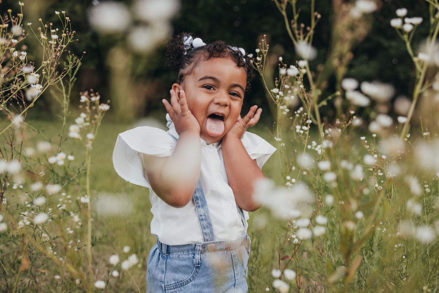 A young child in denim overalls and a white shirt makes a playful expression while standing in a wildflower meadow.