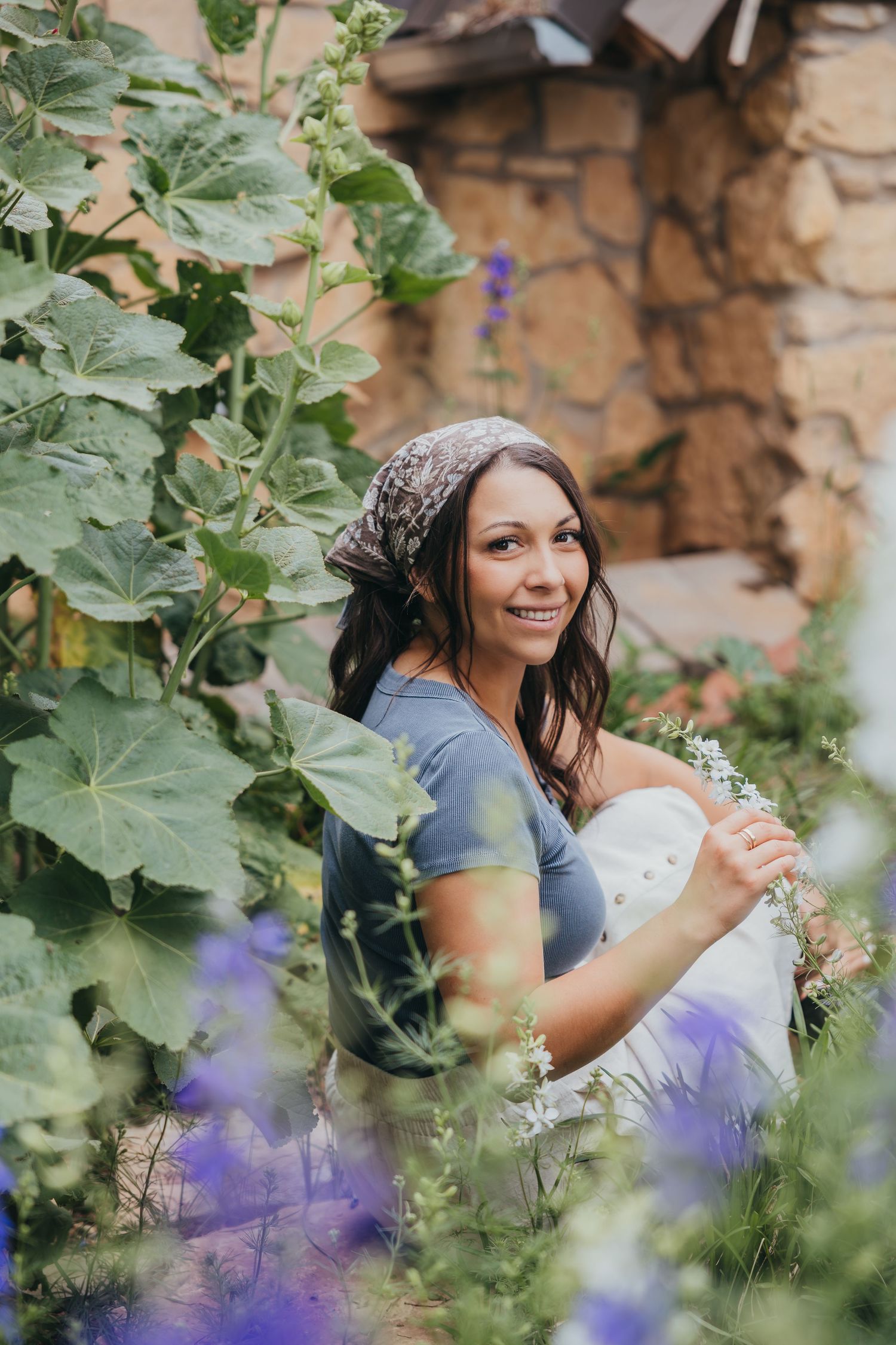 A person in a blue top sits among purple flowers and lush greenery in a garden setting with a stone wall background.