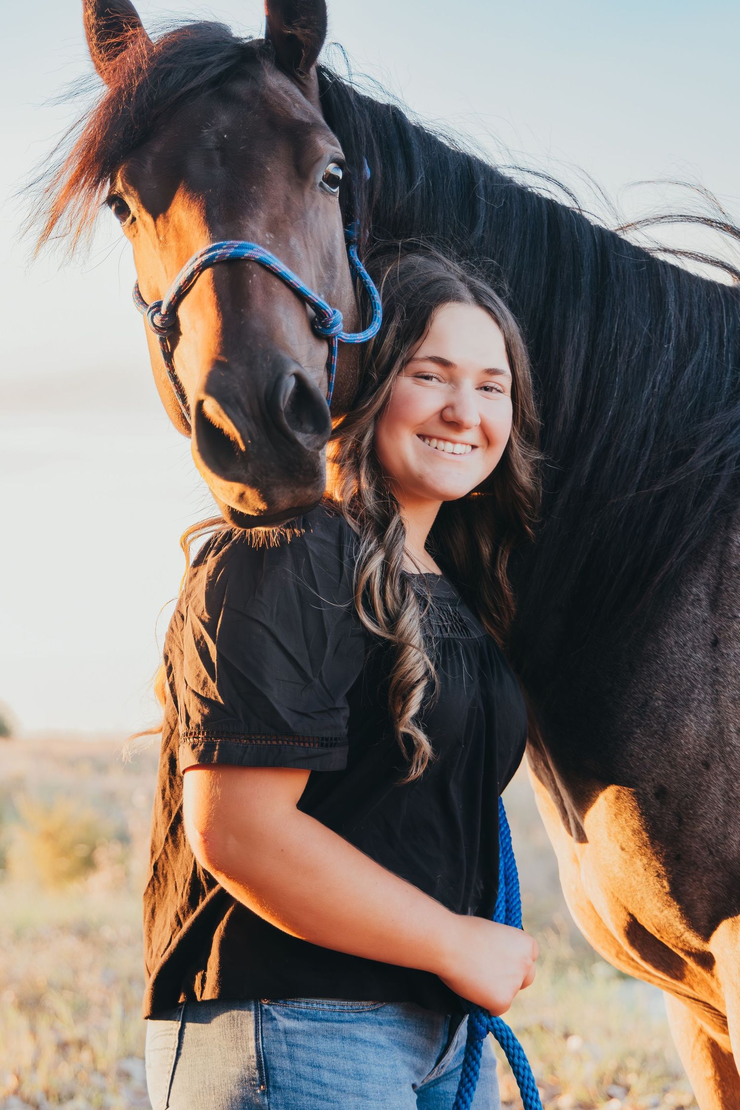 Young woman smiling and embracing a dark brown horse with a blue halter outdoors in a sunny field.