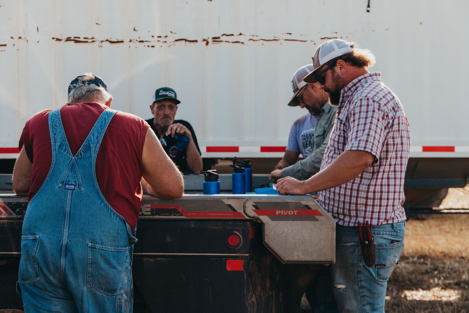 Agricultural workers in overalls and casual wear gathered around the bed of a pickup truck discussing work tasks.