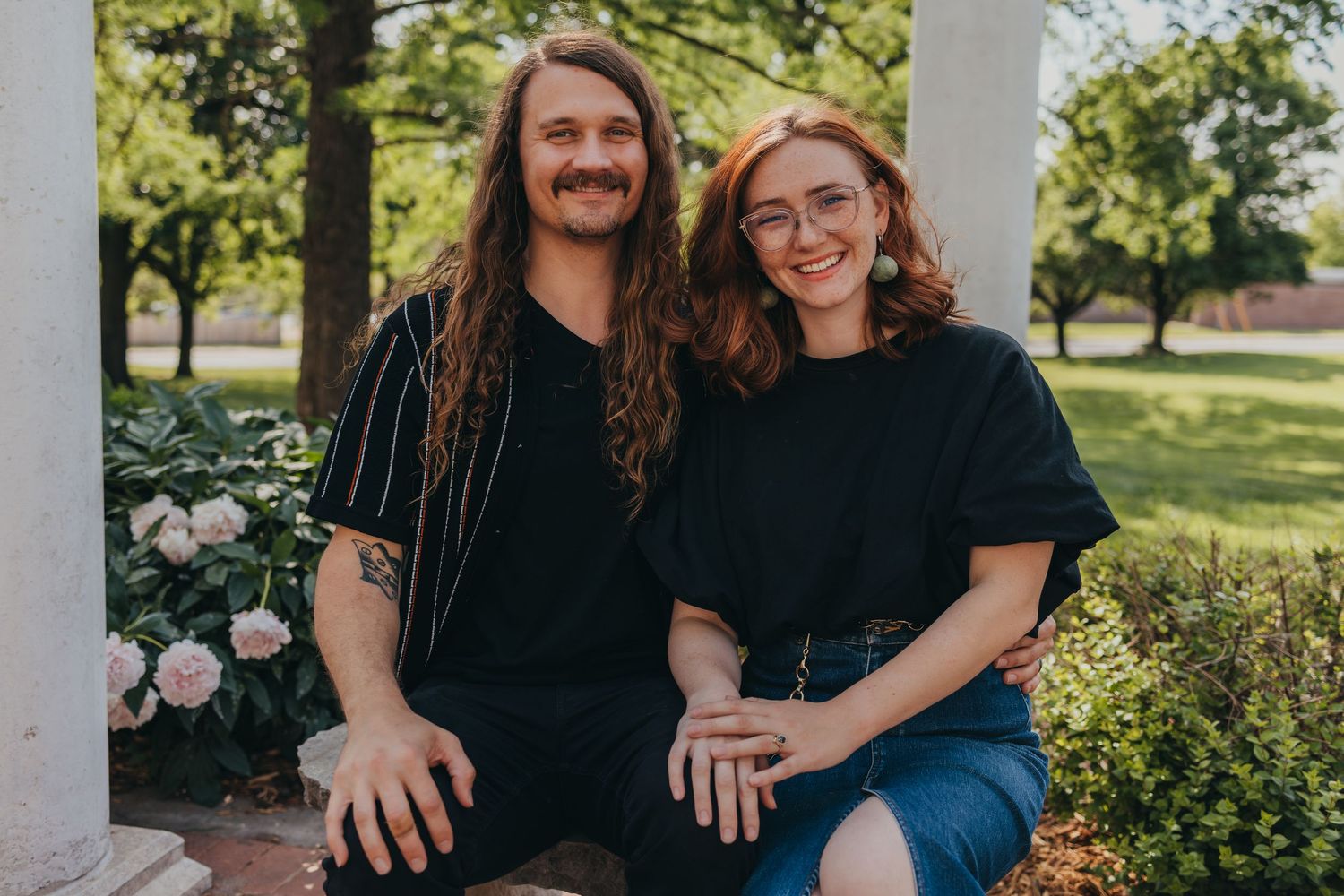 Two people in black shirts sit together on outdoor steps surrounded by blooming flowers and greenery in a garden setting.