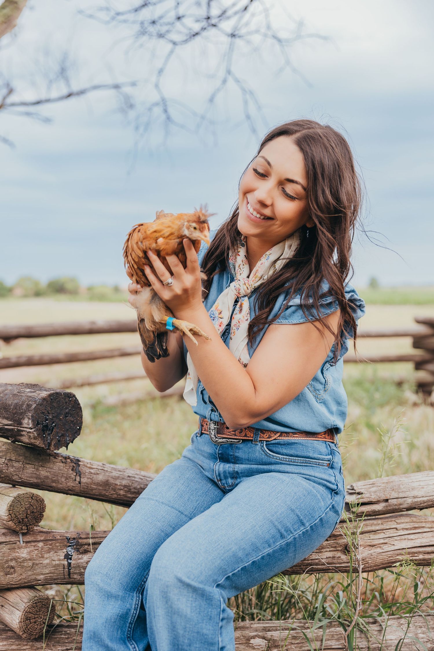 A woman in denim sits on wooden logs holding a brown chicken while enjoying a sunny day in a rural setting.