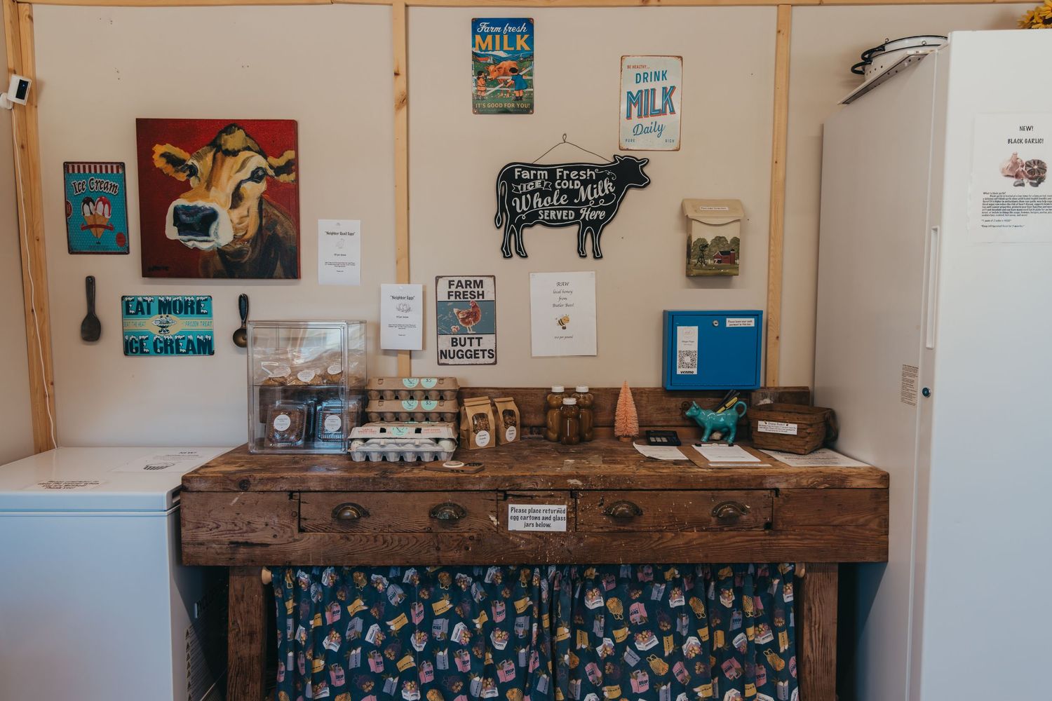 Rustic farmhouse kitchen display with wooden table, floral curtains, and vintage cow-themed artwork on cream colored walls.