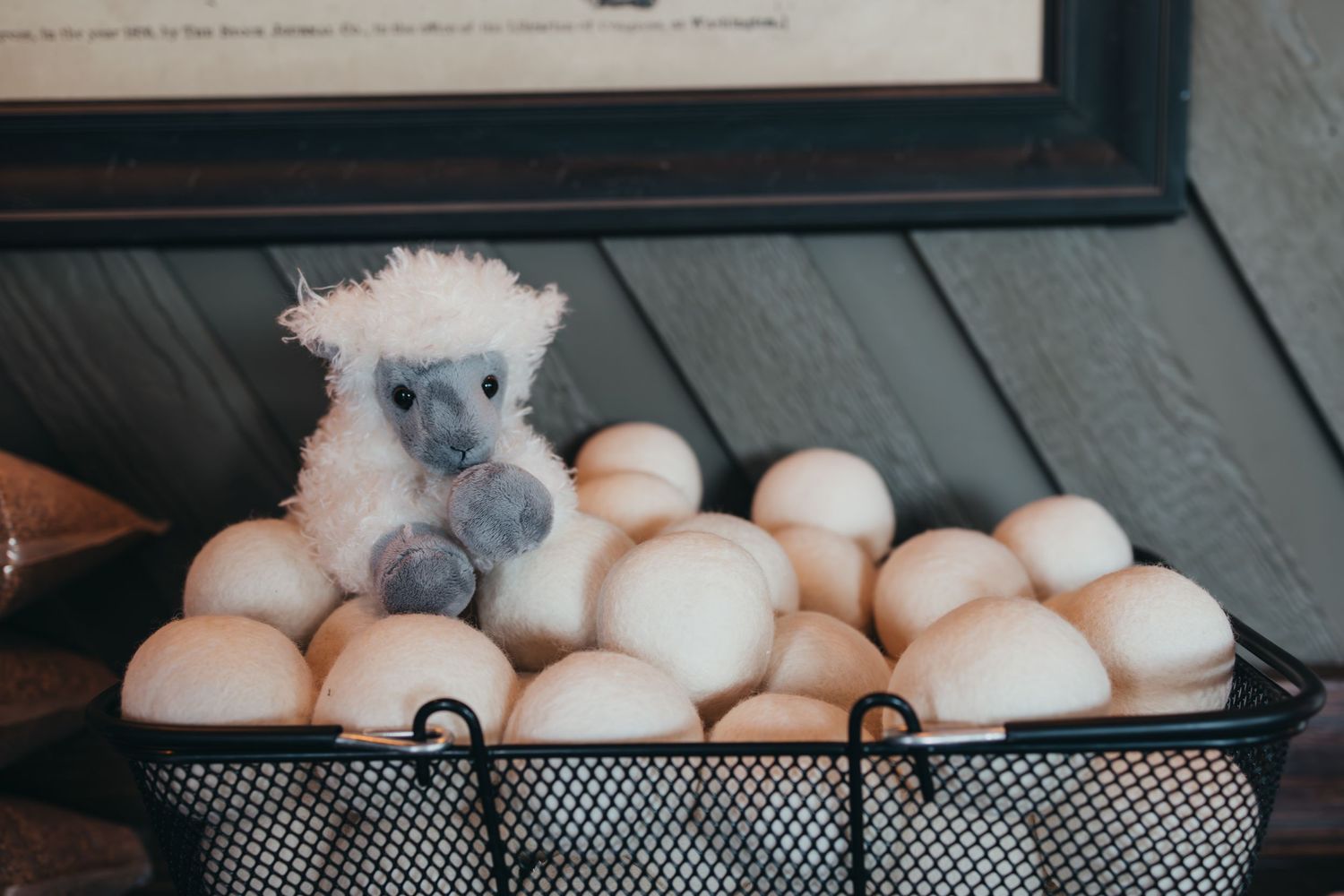 A small grey stuffed animal sits in a black wire basket filled with fresh white eggs.