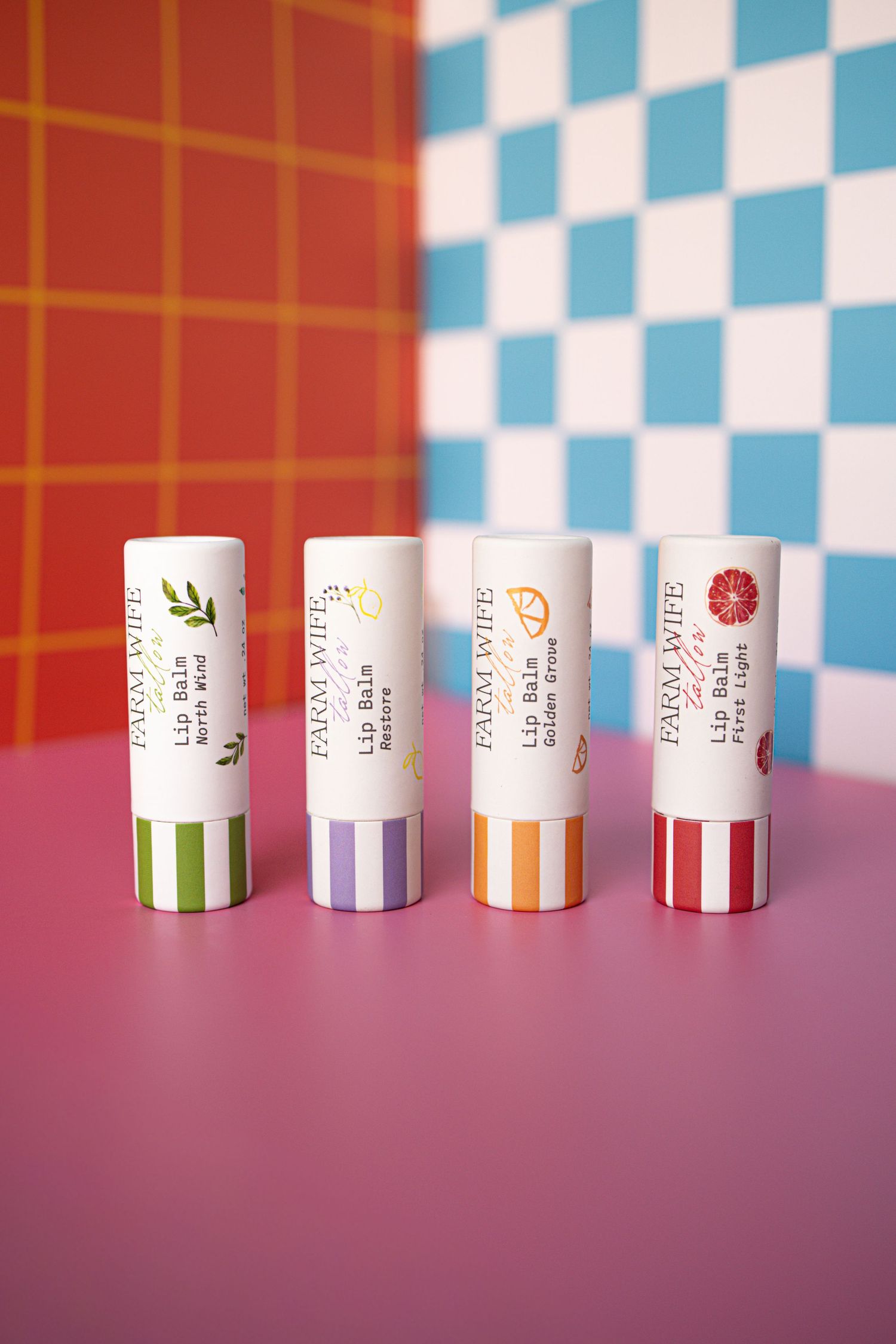 Four Farm Wife lip balms in assorted flavors displayed upright on a pink surface with colorful checkered background.