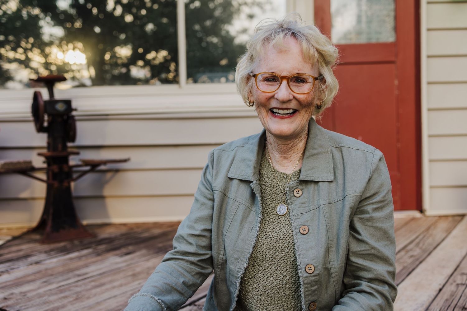 A cheerful person in a gray jacket and glasses sits on a wooden deck during sunset with a warm, genuine smile.