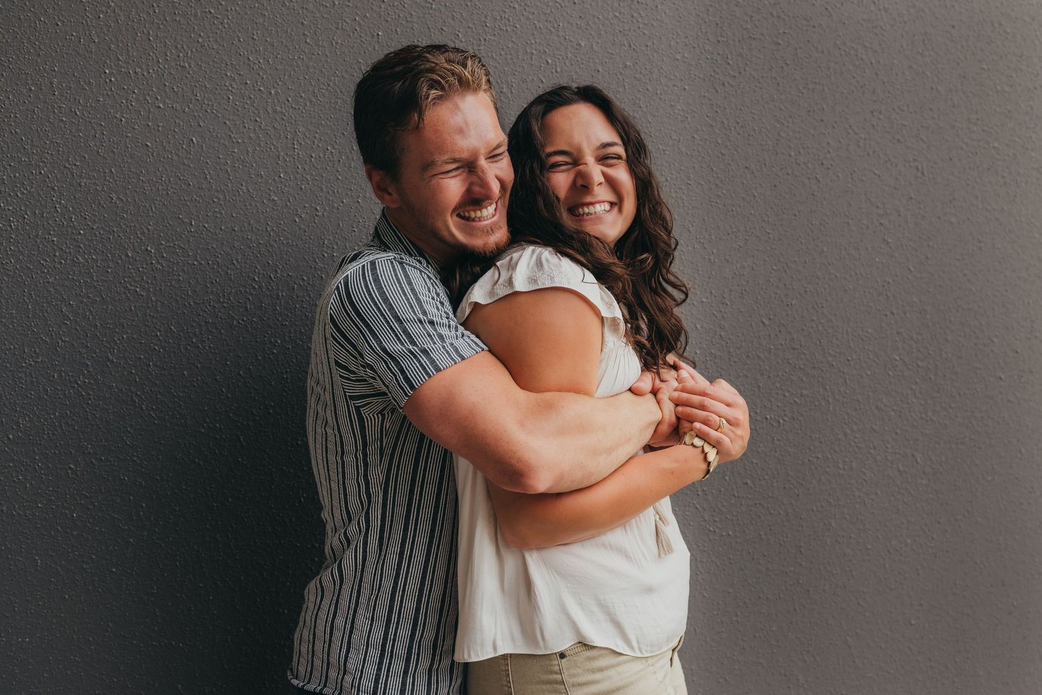 A happy couple shares a tender moment as they embrace and laugh against a gray wall.