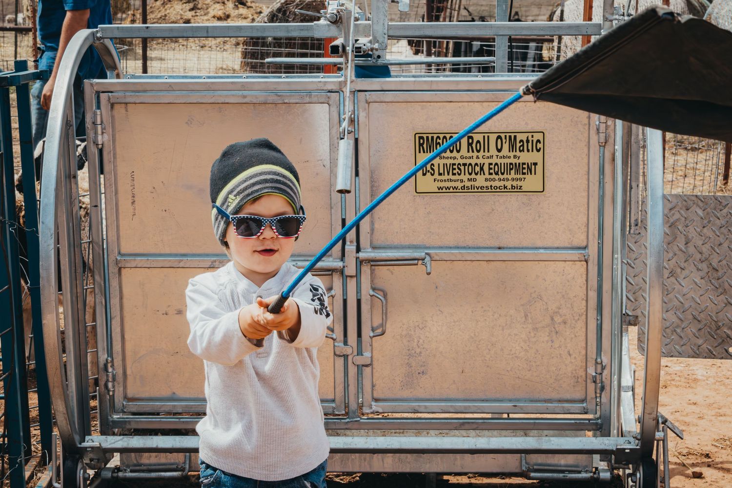Young rancher in white sweater and sunglasses near livestock handling equipment.