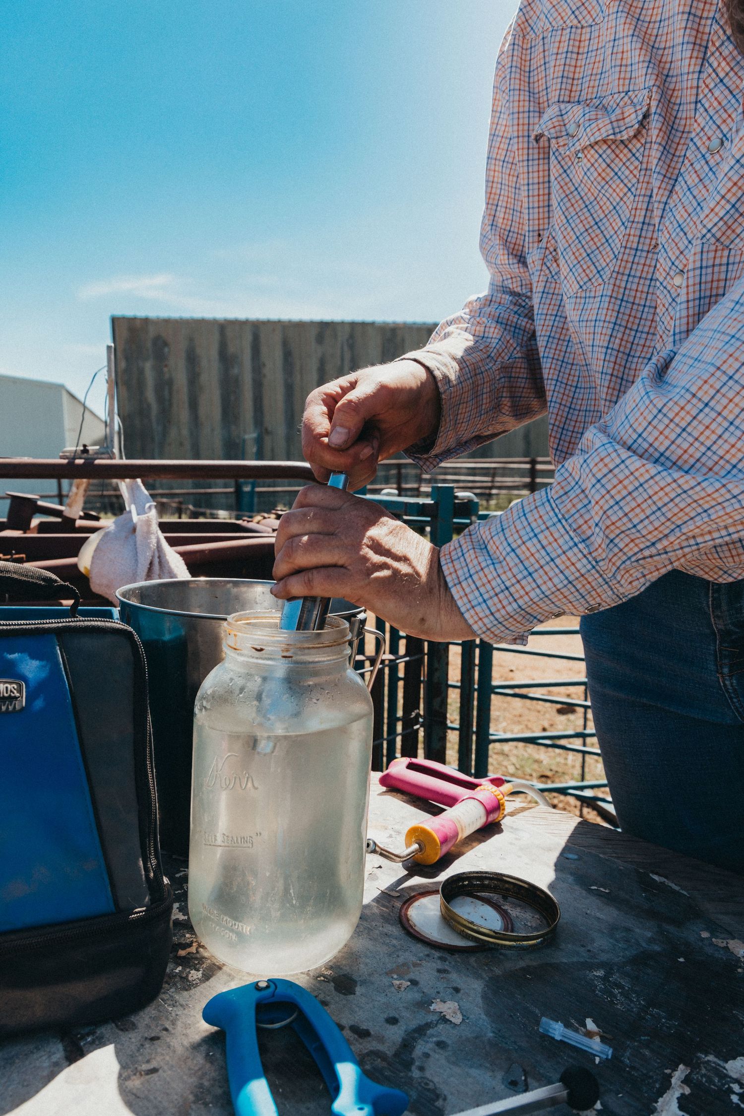 Person in plaid shirt filling glass jar outdoors with tool supplies nearby.