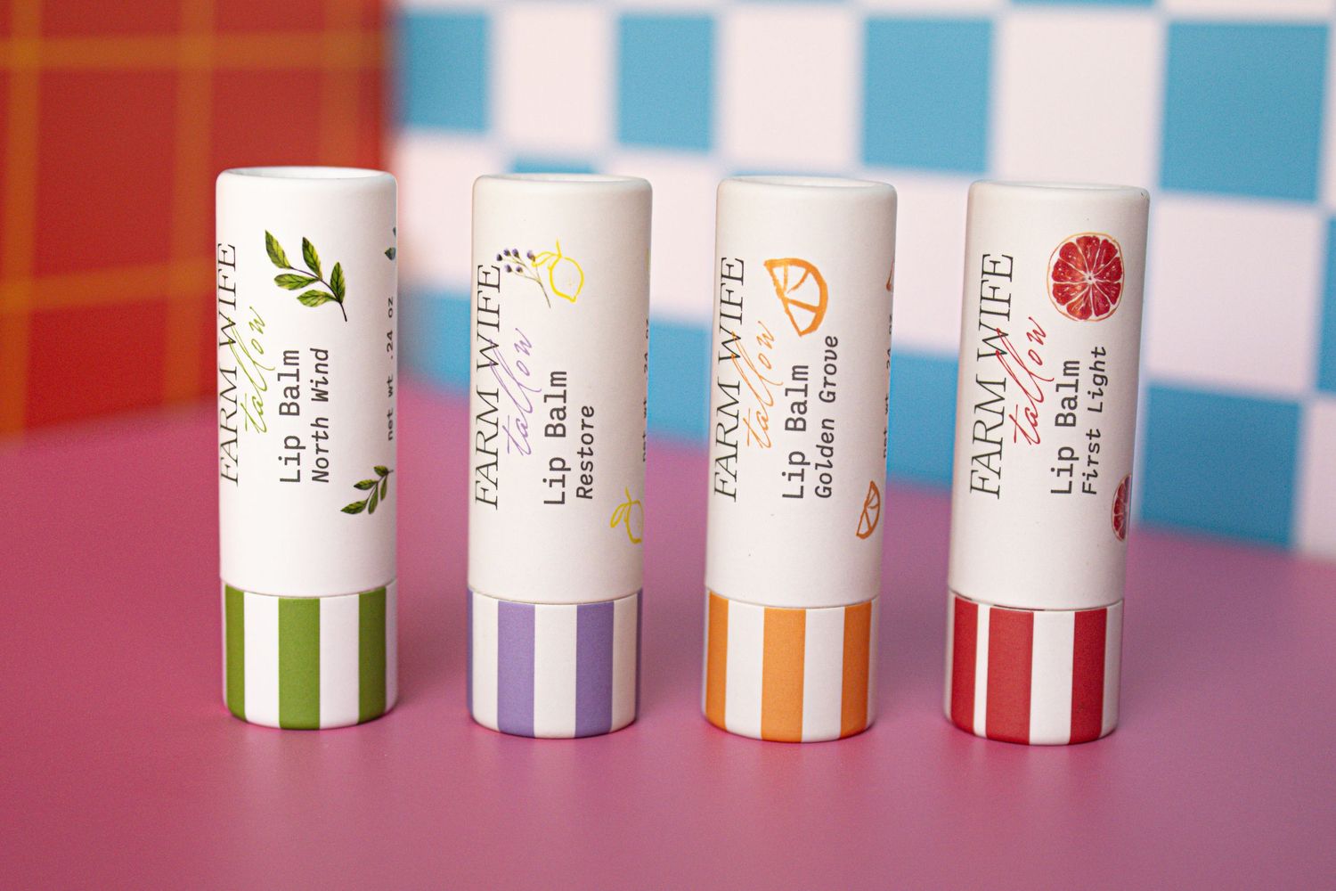 Four Farm Wife lip balm sticks in colorful striped packaging displayed on a pink surface with blue tile background.