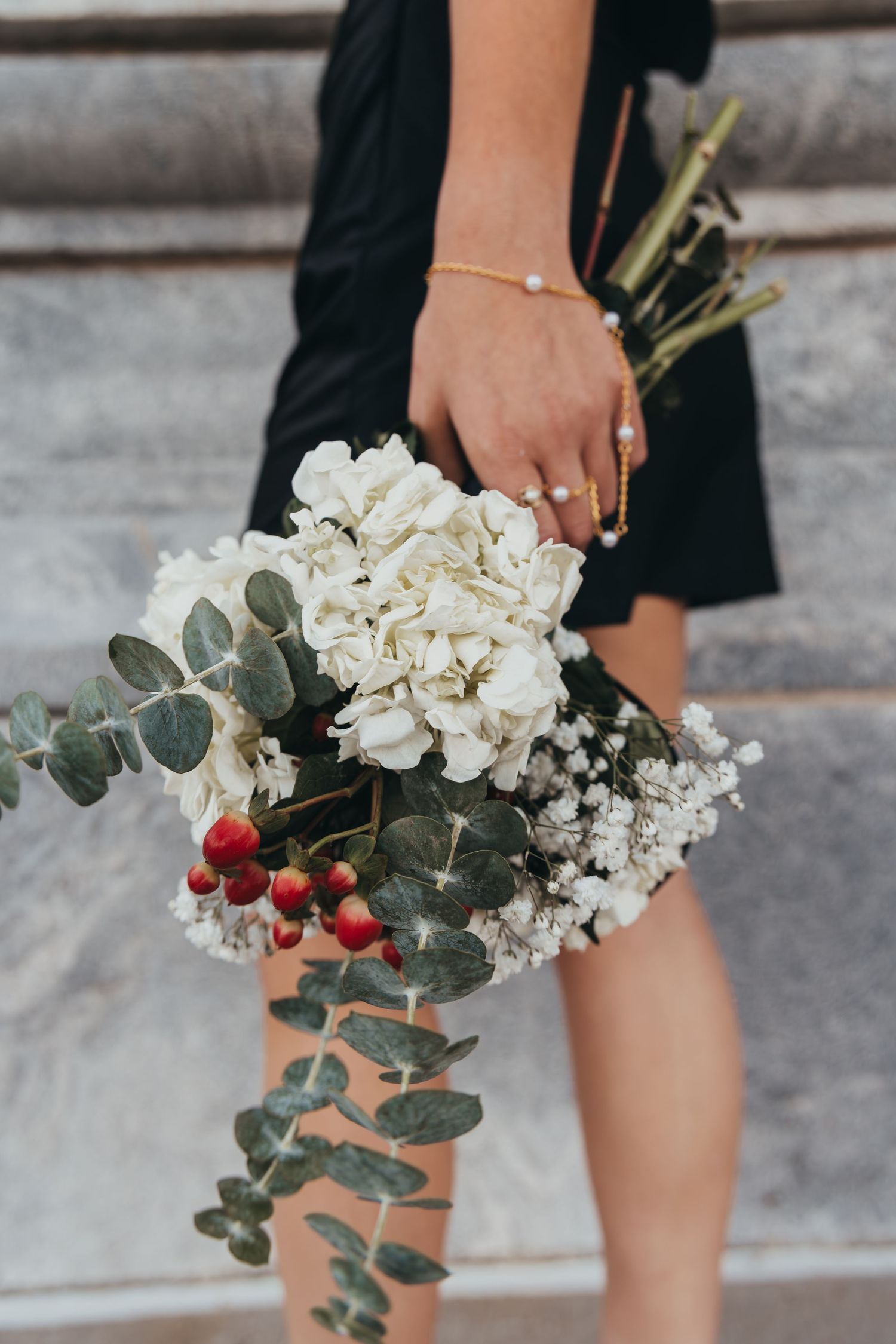 A bouquet of white flowers and eucalyptus leaves held against a dark dress and concrete background.