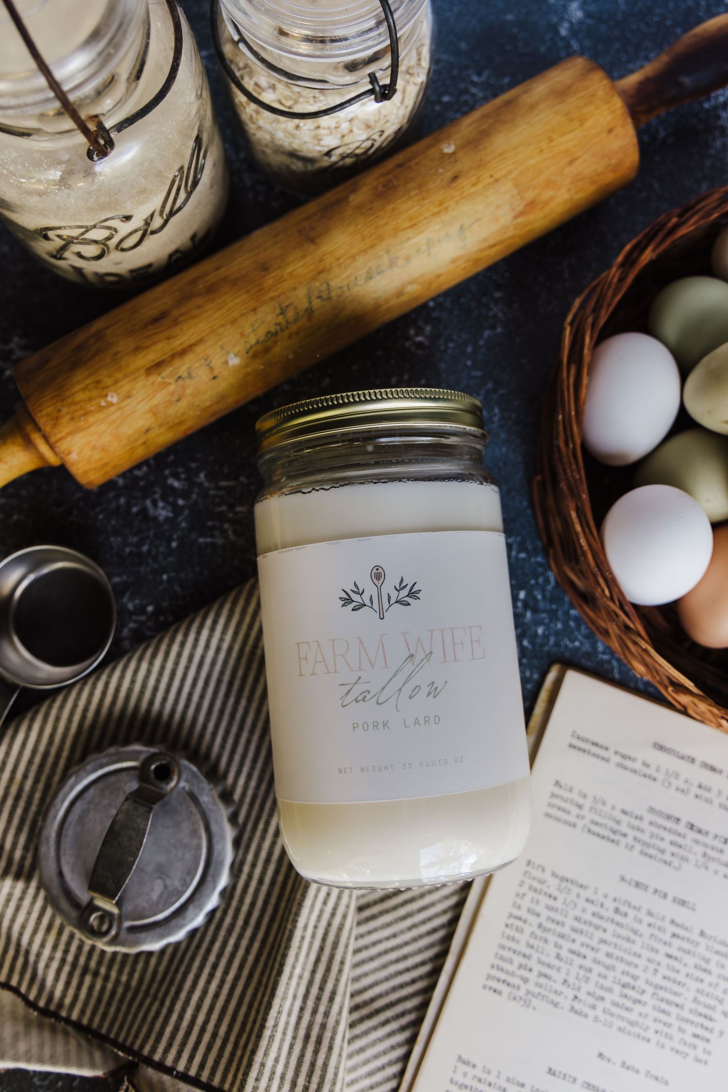 Vintage mason jars, eggs, and a wooden rolling pin arranged with handwritten recipe cards on a dark rustic surface.