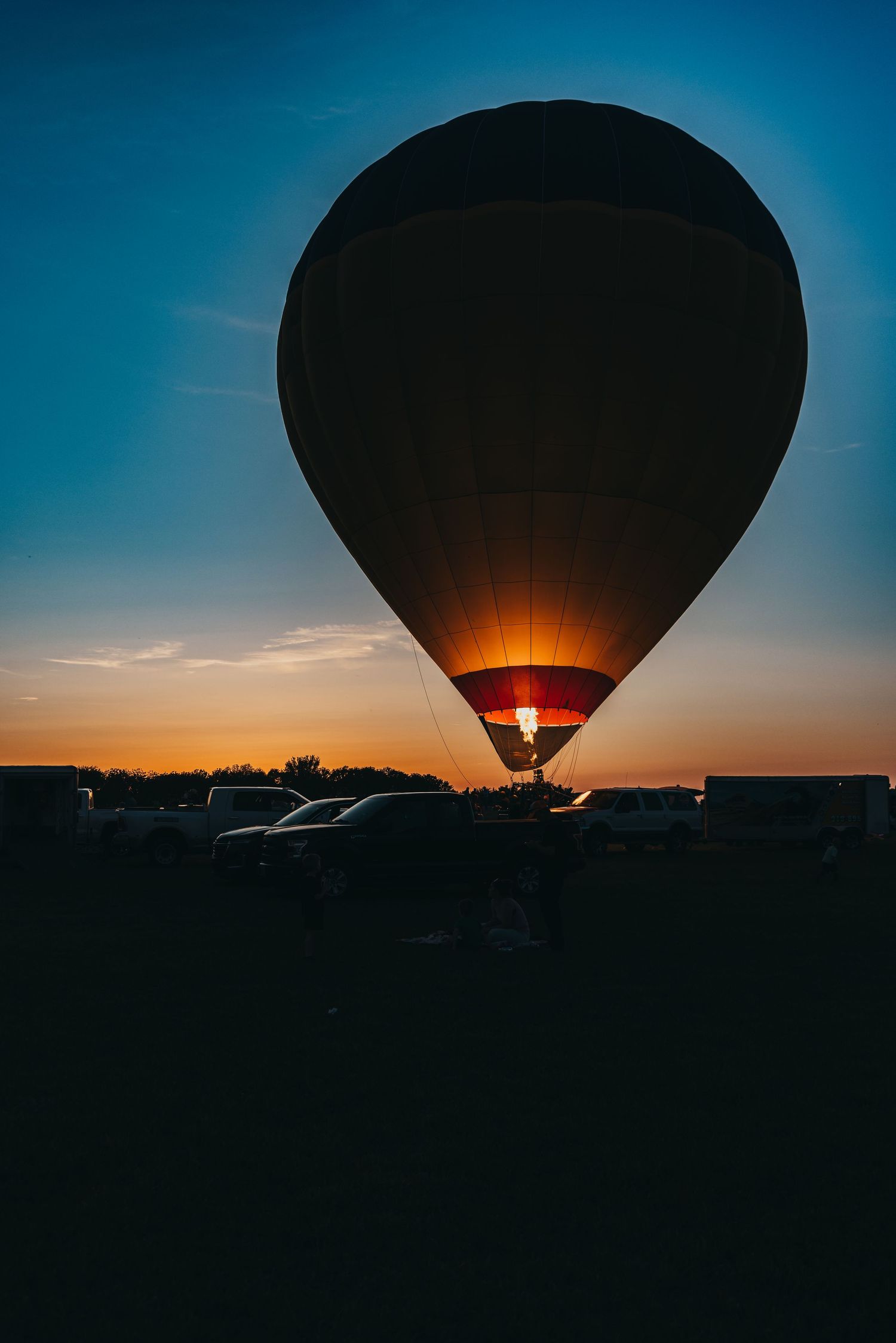 Silhouette of hot air balloon glowing against sunset sky with parked cars below.