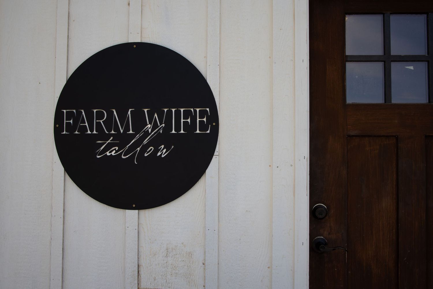 Black circular Farm Wife sign mounted on white wooden exterior wall.