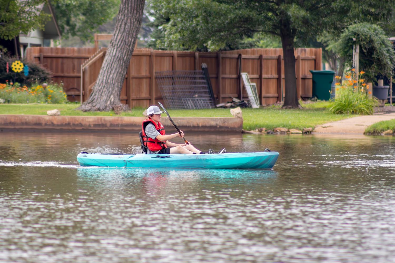 A person in red paddling a turquoise kayak along the shoreline of a peaceful lake.
