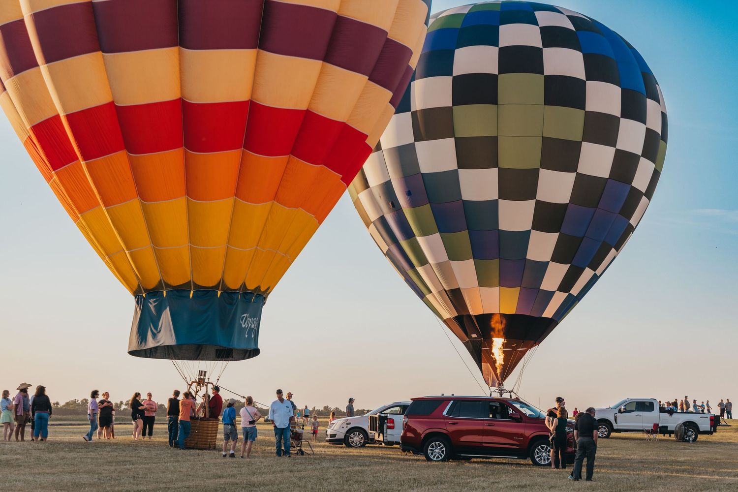 Two hot air balloons, striped and checkered, prepare for launch as spectators gather.