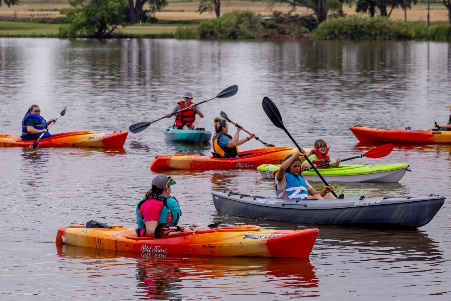 Colorful kayakers paddling across a calm lake with trees in background.