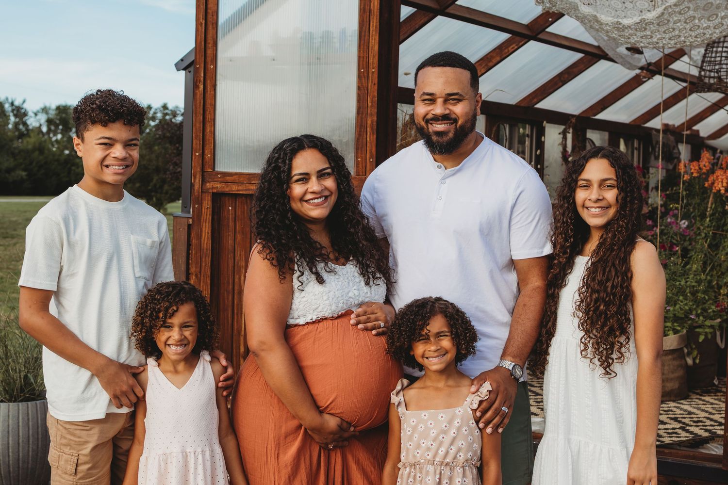 Family of six poses outdoors near greenhouse wearing coordinated neutral and rust-colored outfits.