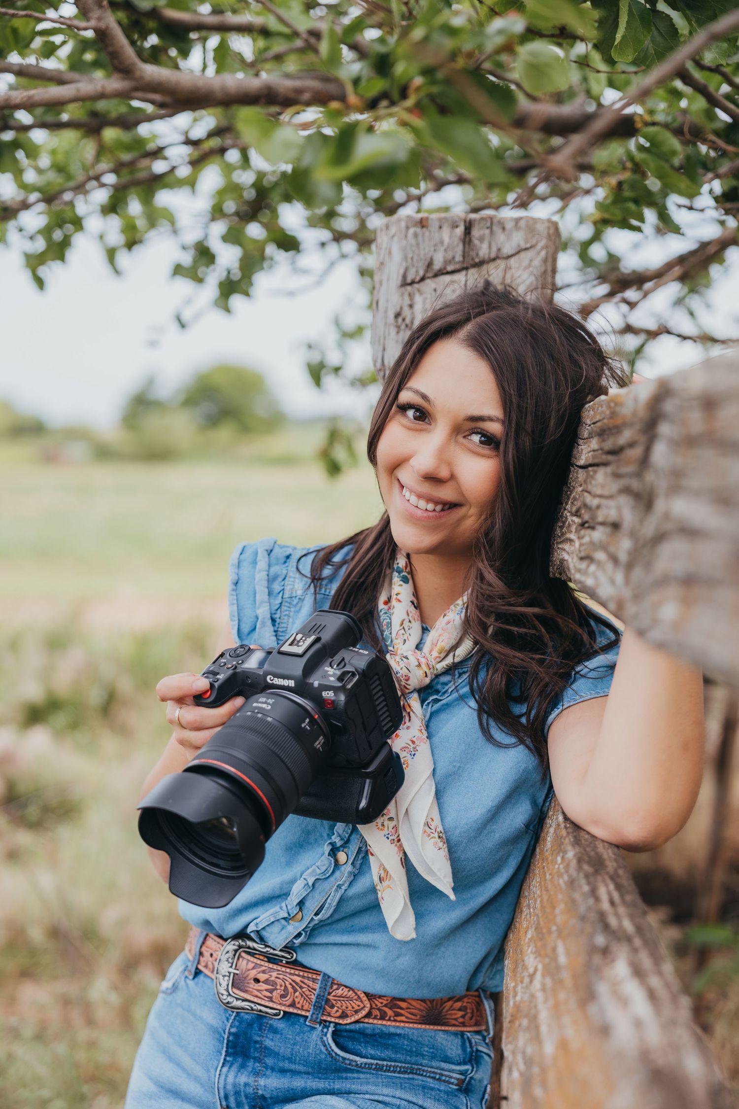 Photographer in denim vest holding professional camera with telephoto lens leans against tree in outdoor setting.