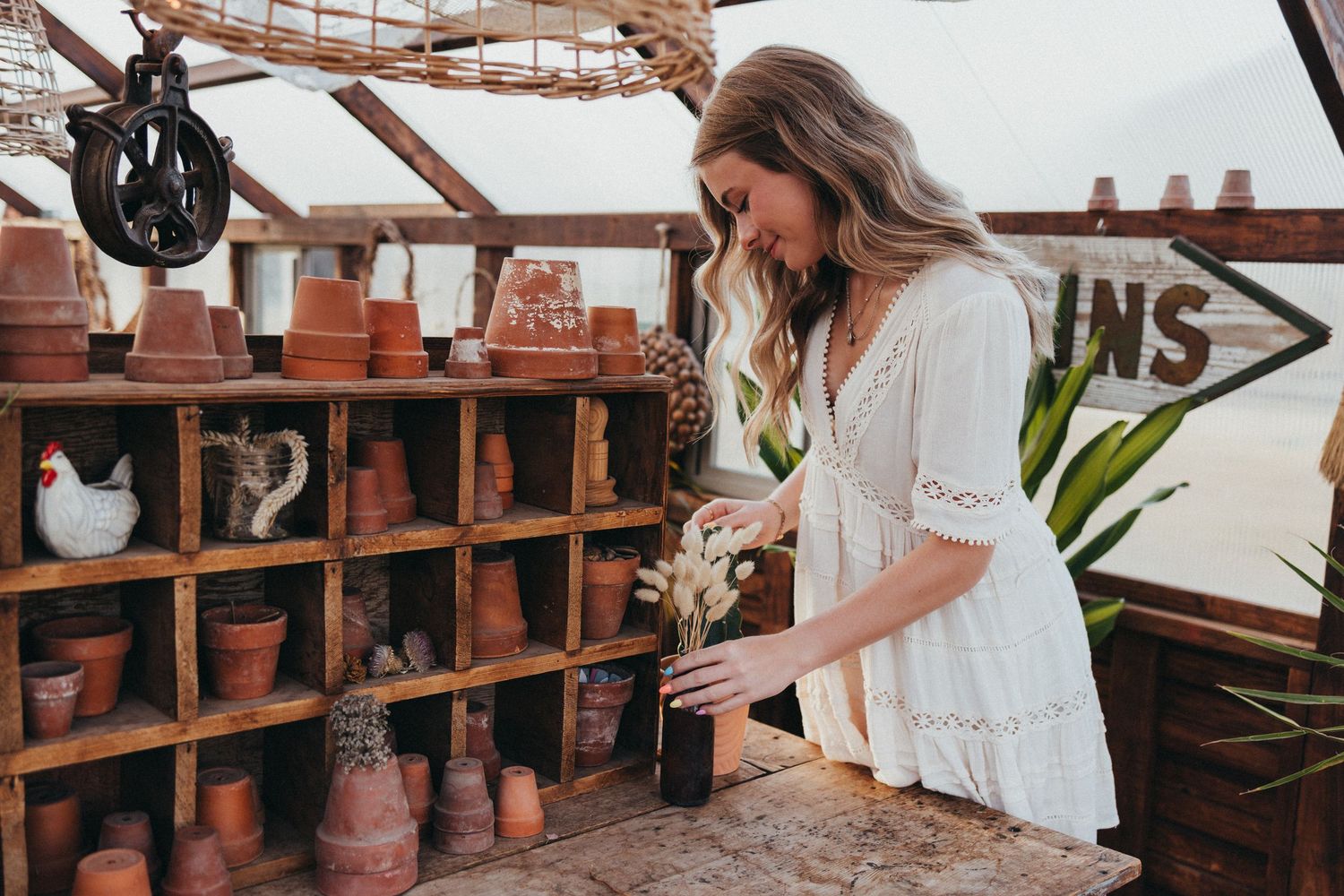 Person in white lace dress arranging terracotta pots on wooden shelving unit in rustic greenhouse setting.