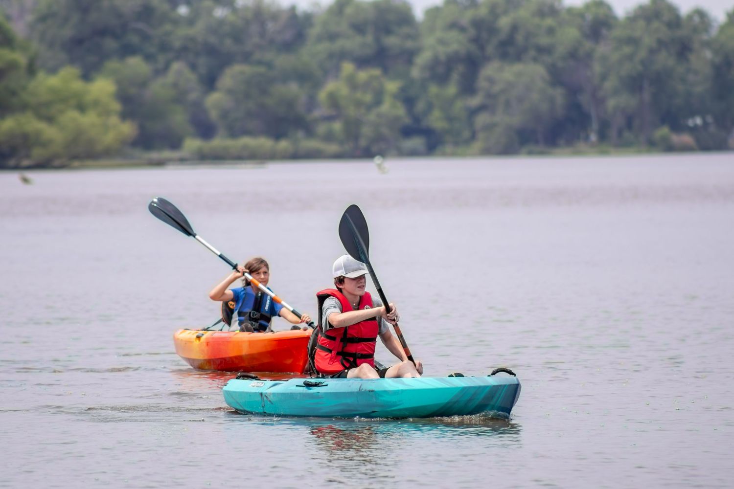 Two kayakers paddle colorful orange and turquoise kayaks across a calm lake surrounded by trees.