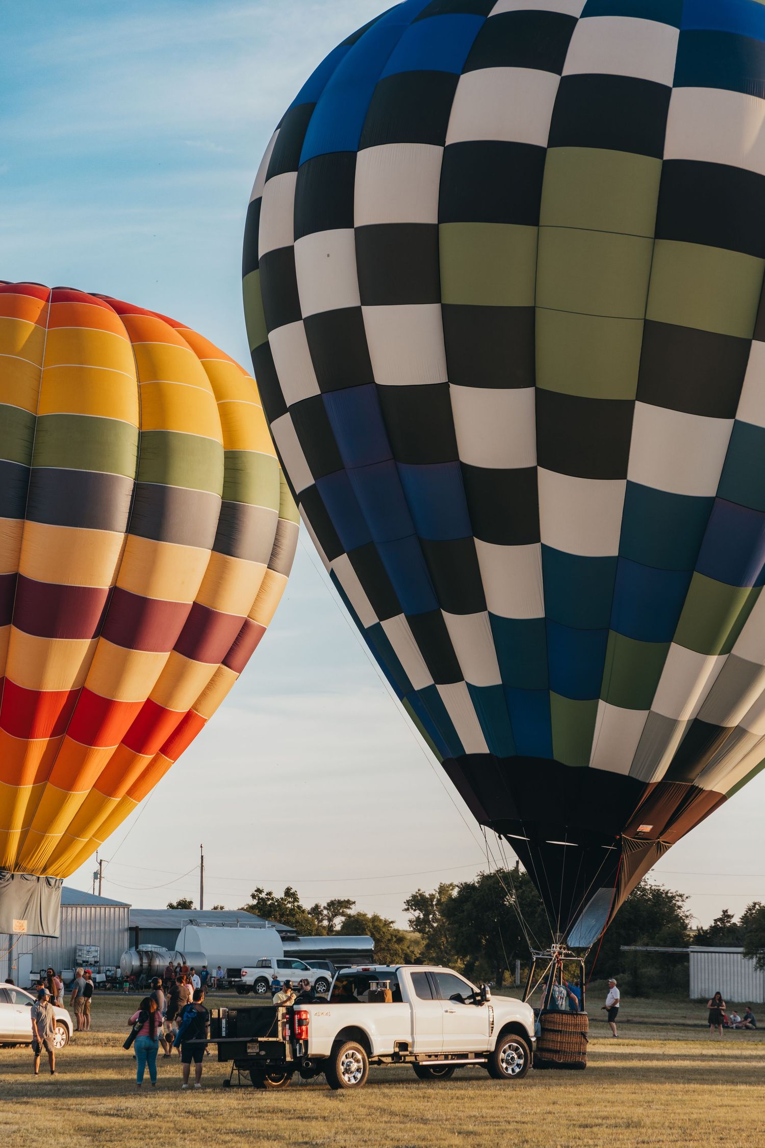 Two vibrant hot air balloons, one checkered and one striped, prepare for takeoff in a field.
