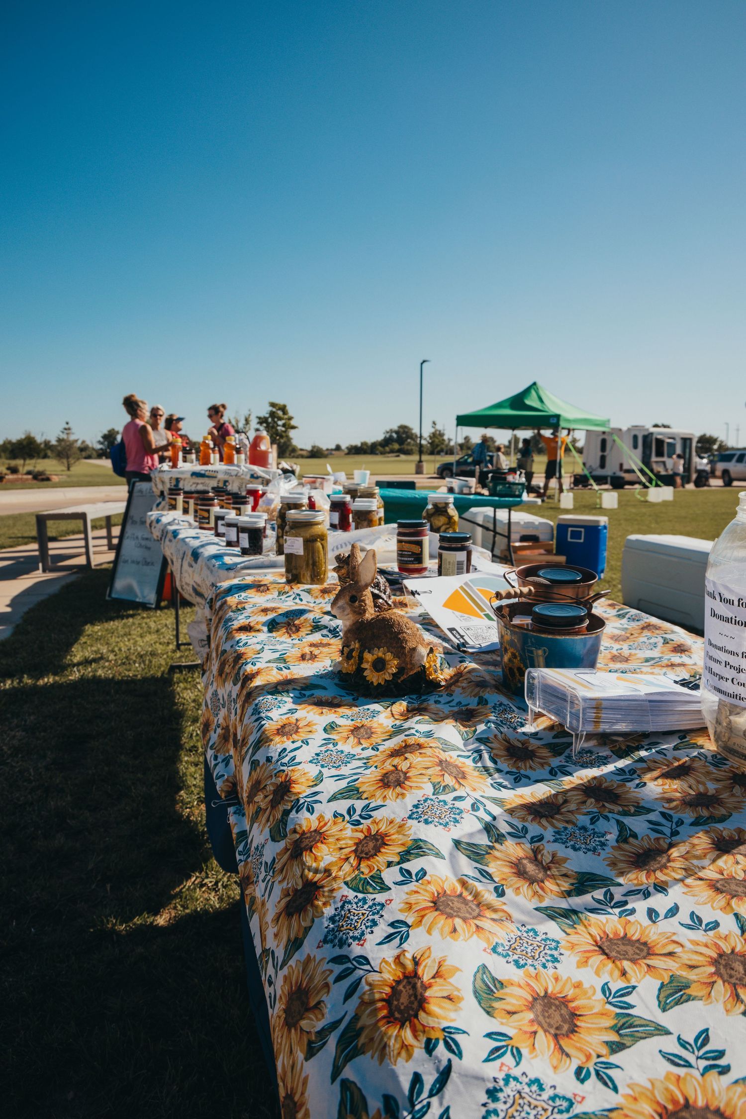 Outdoor market stalls with sunflower tablecloths and canopies set up in a grassy field on a sunny day.