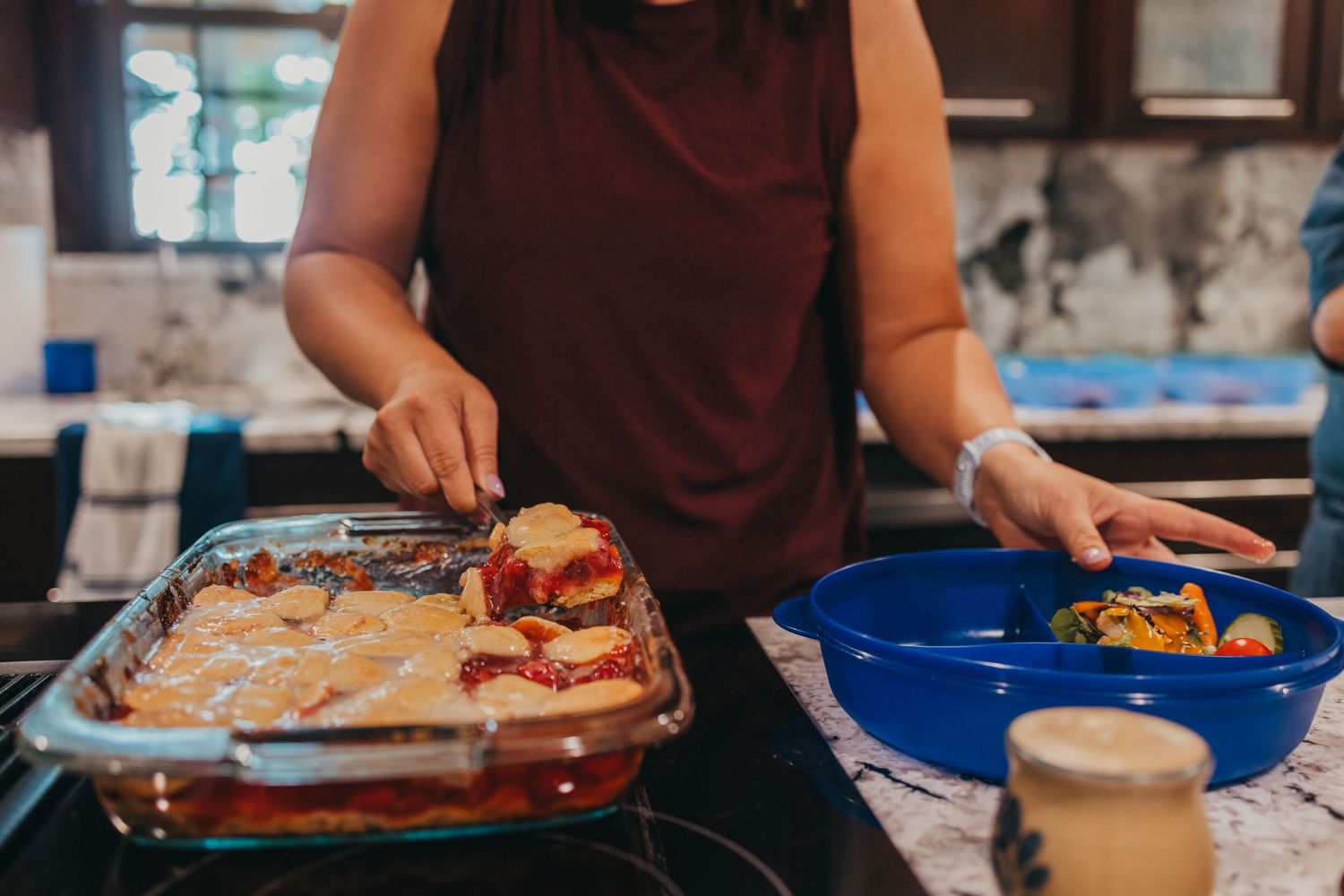 Person in maroon shirt serving food from a casserole dish onto blue plates in a kitchen setting.