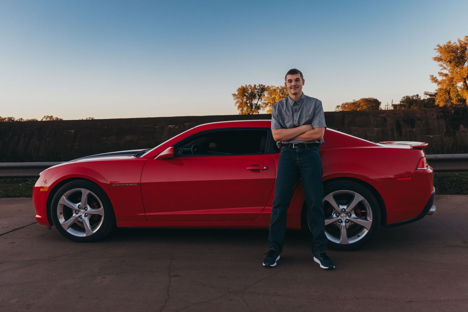 Man posing with arms crossed beside a red Chevrolet Camaro at sunset on an open road.