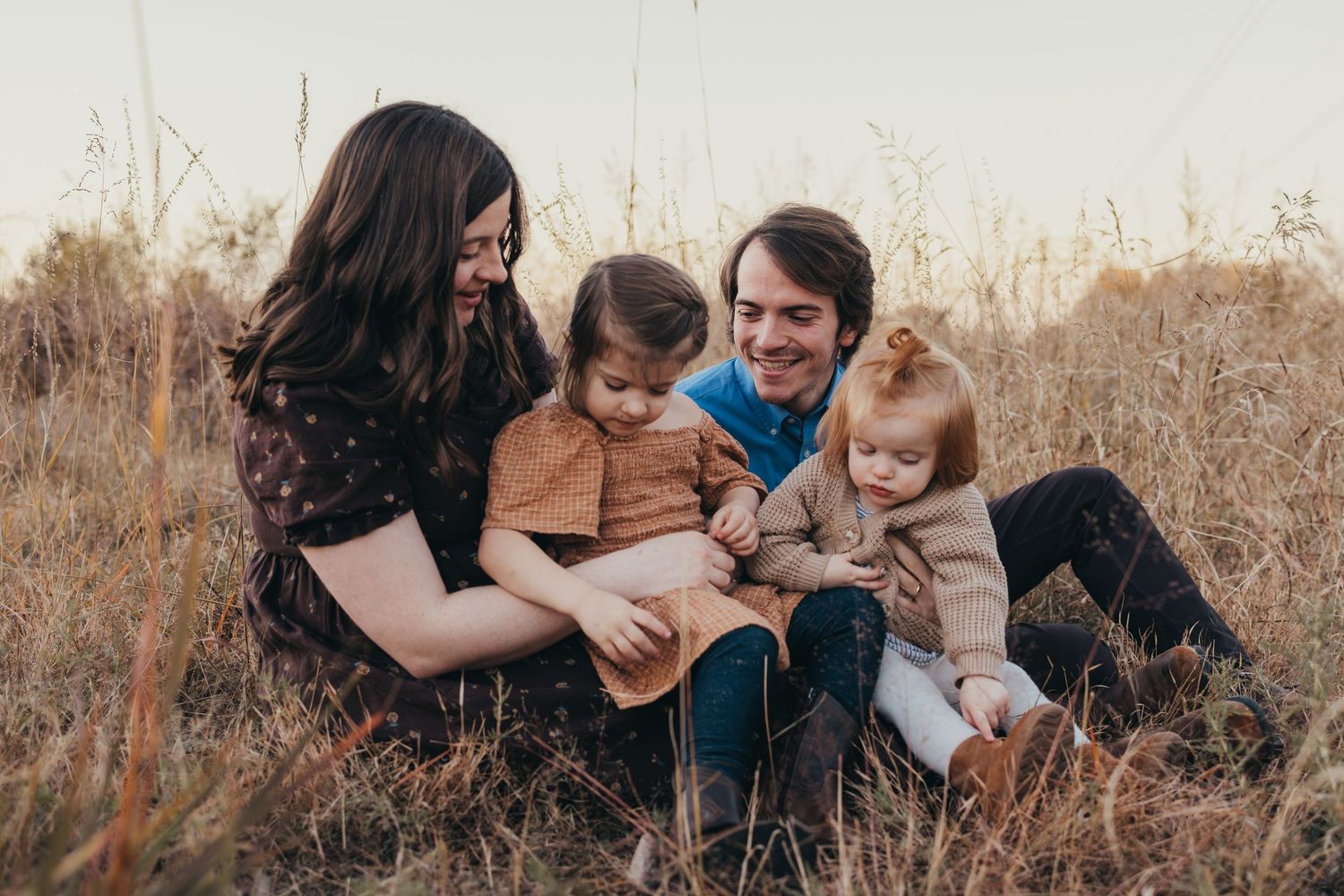 Family of four sitting together in a golden field, parents smiling at two young children in warm autumn tones.