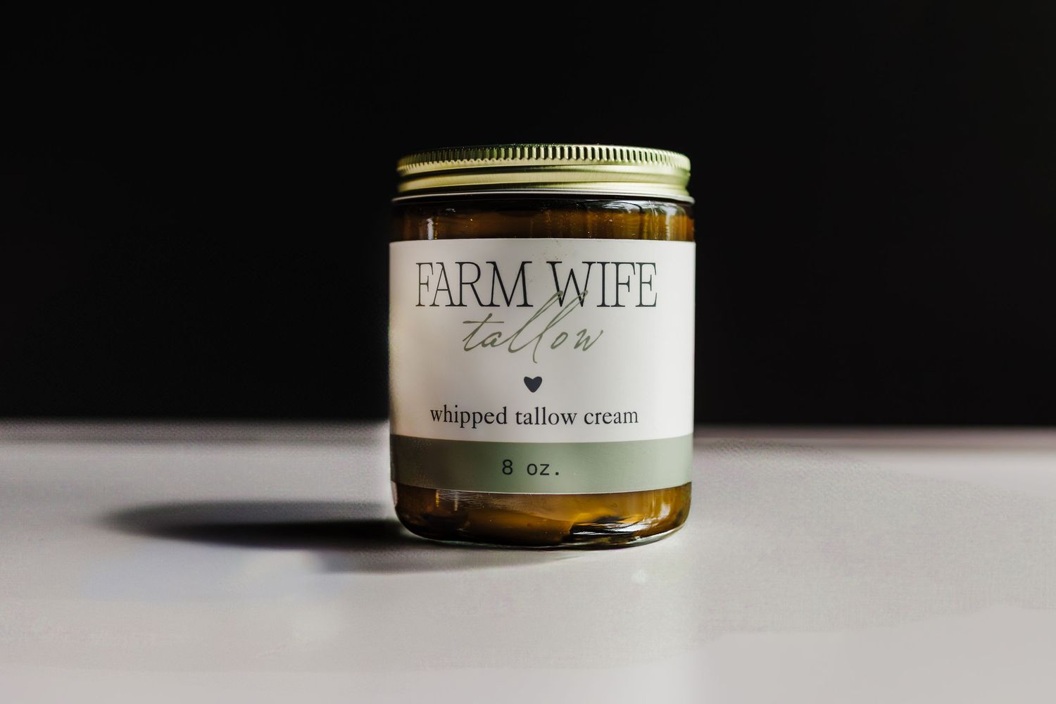 A glass jar of Farm Wife Collective whipped tallow cream sits on a white surface against a black background.