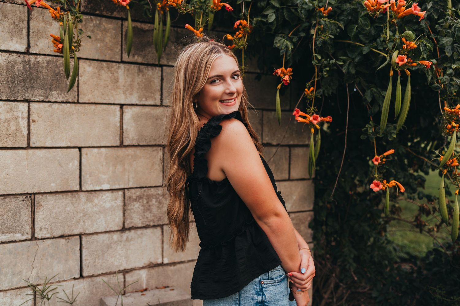 Someone stands by a stone wall with blooming orange flowers and green foliage in the background.