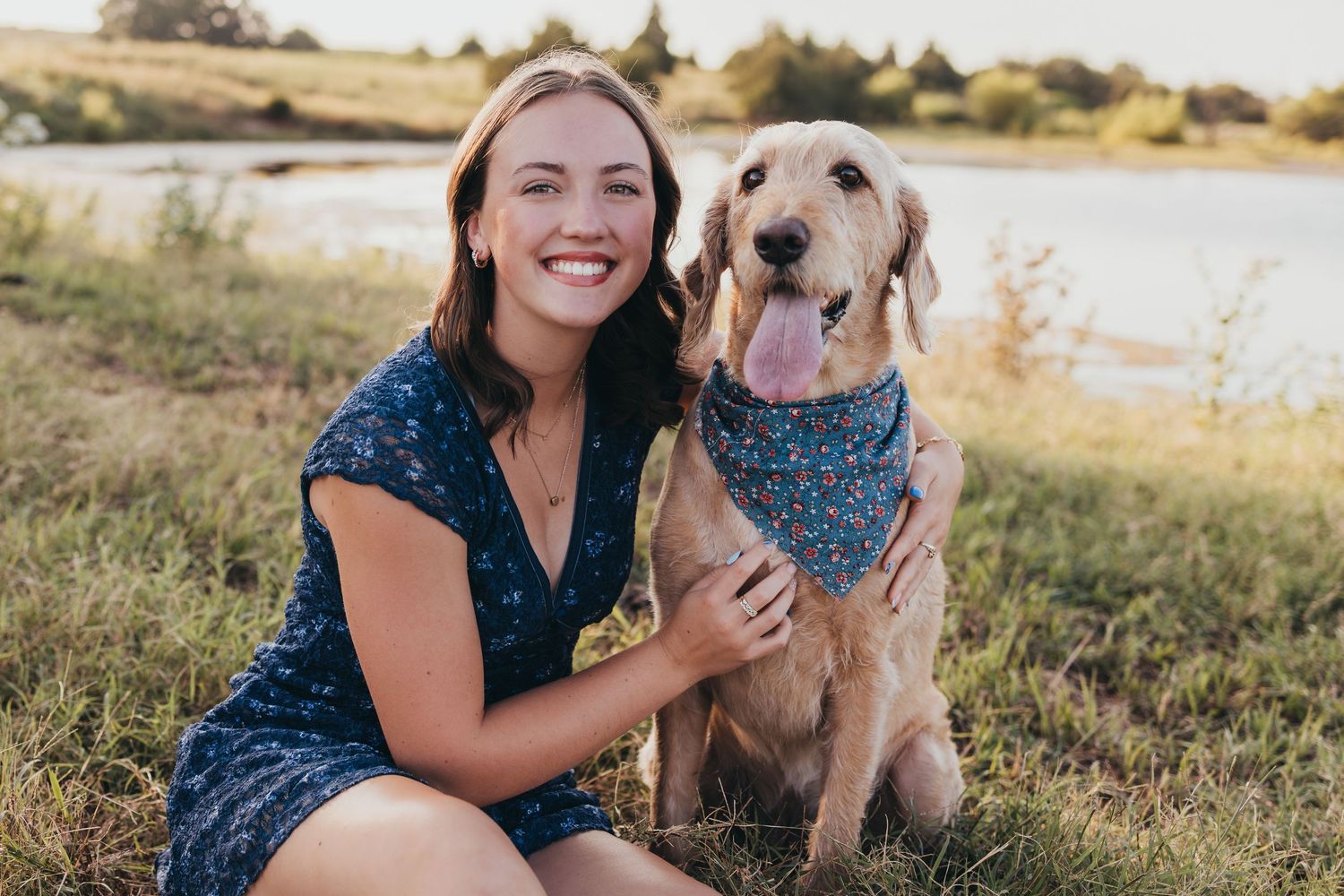 A happy golden retriever dog wearing a blue bandana plays outdoors with their companion in a sunny field at sunset.