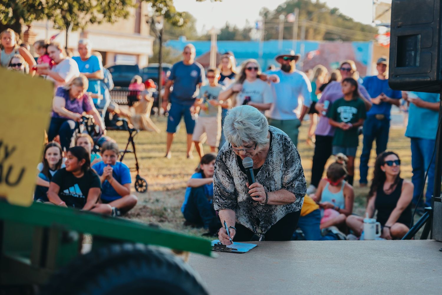 A crowd watches an outdoor performance with spectators sitting on the ground at dusk.