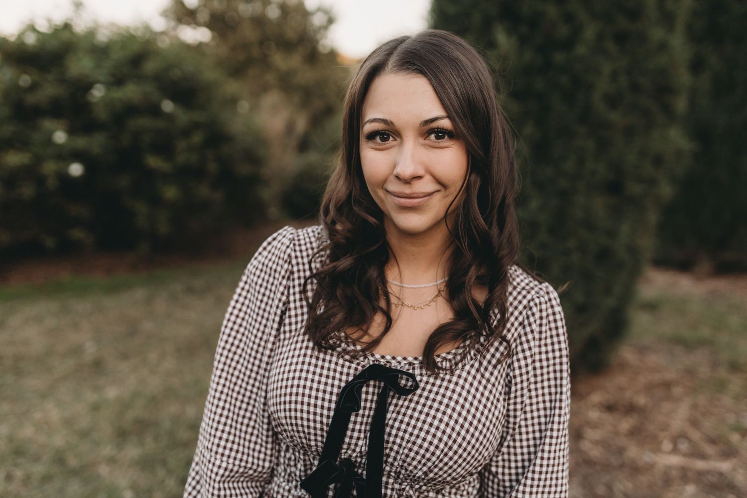 Woman in black and white checkered dress poses outdoors in a natural setting during golden hour.