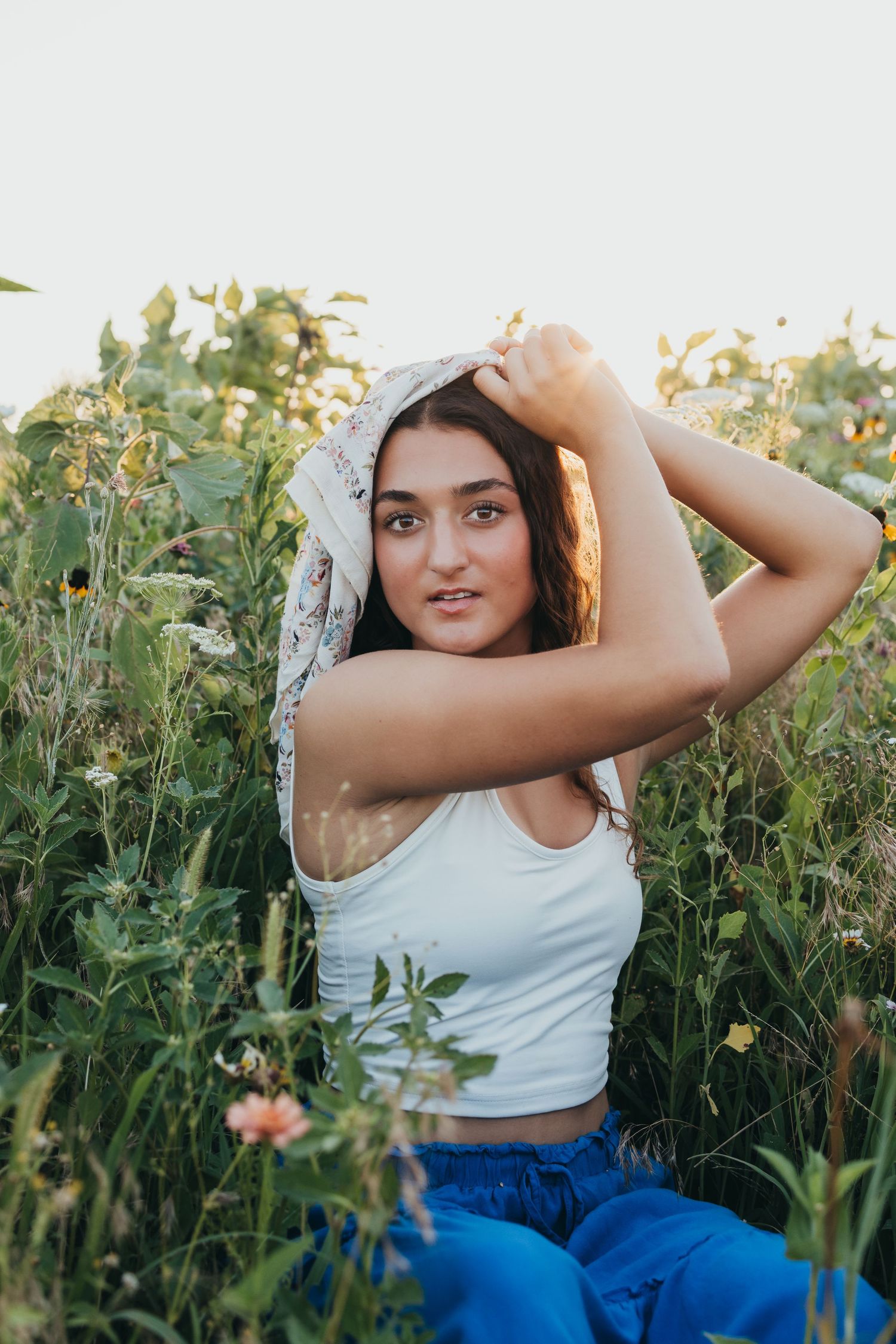A woman in a white tank top poses gracefully among wildflowers during golden hour.