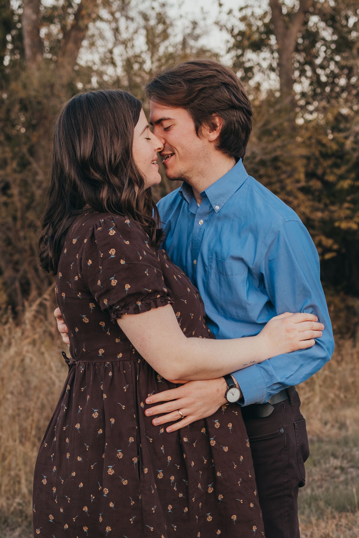 A couple shares an intimate moment outdoors in autumn, with her in a brown dress and him in a blue button-down shirt.