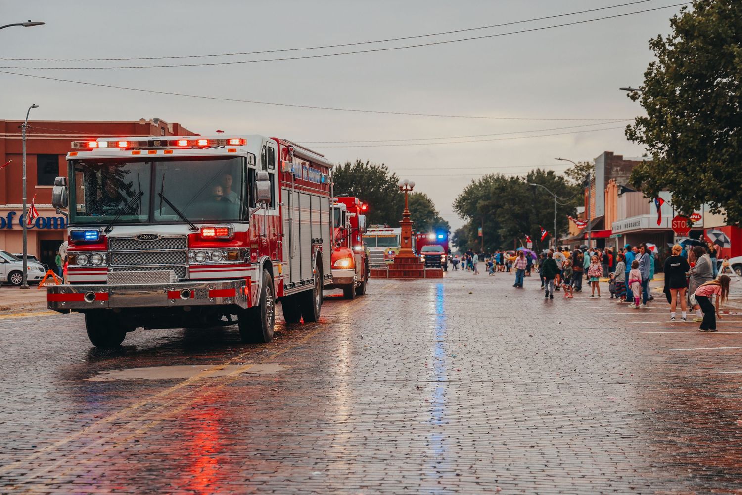 Fire trucks parading down wet brick street during community event.