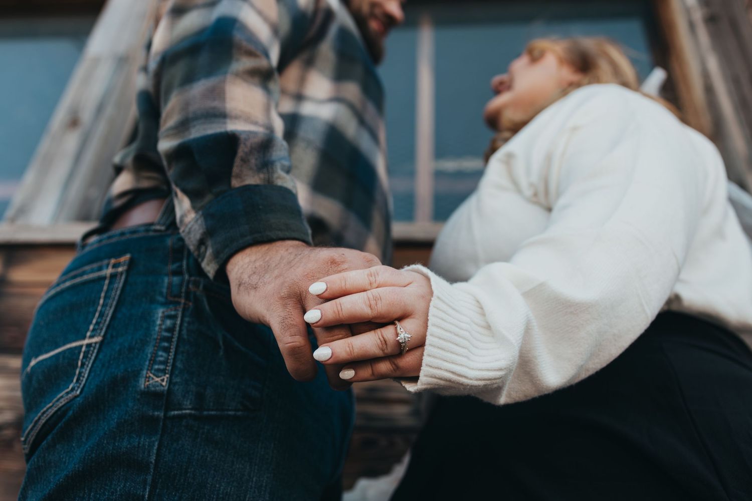 Couple holding hands tenderly, woman wearing engagement ring, man in plaid shirt and jeans, romantic outdoor setting.