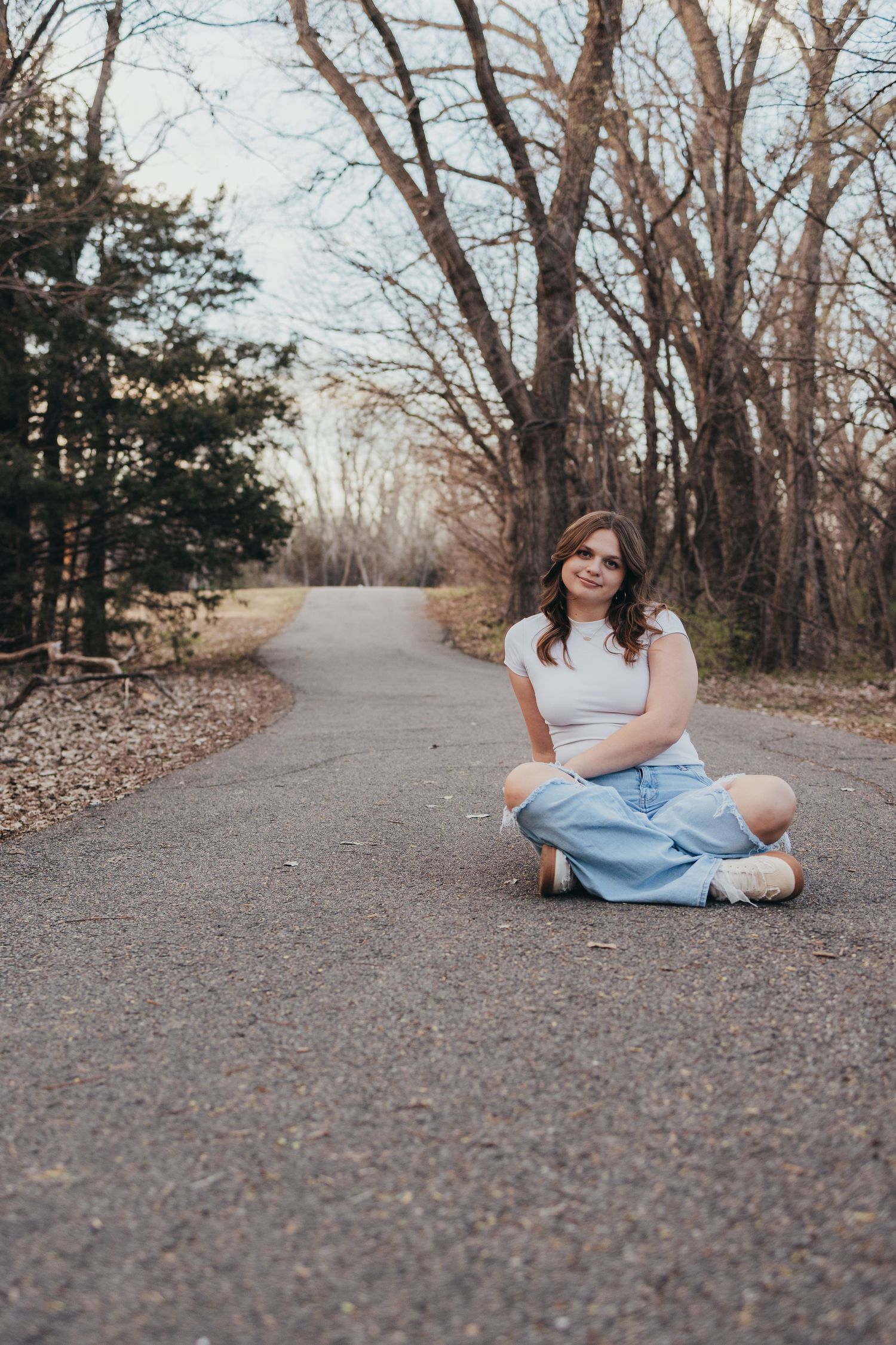 Young woman in white top and blue skirt sitting cross-legged on a winding path surrounded by bare trees.