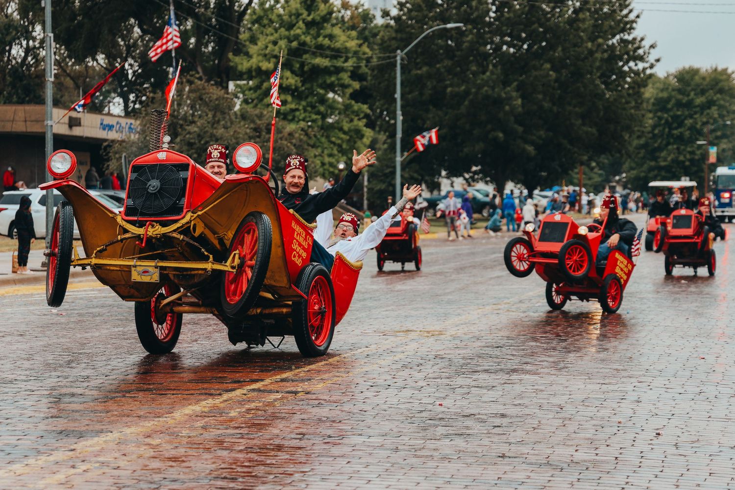 Vintage red cars parade down a wet brick street during a small town celebration.